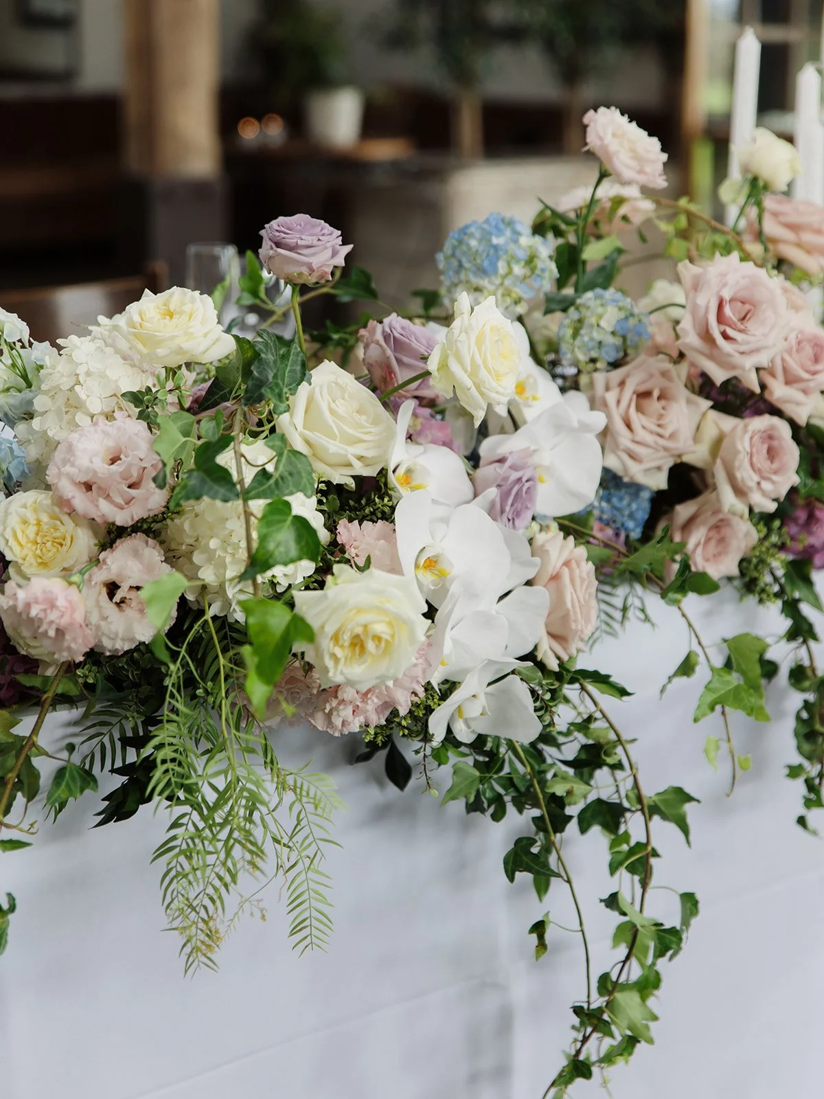 A large floral arrangement featuring white, pale pink, purple roses, white orchids, blue hydrangeas, and green leafy accents, displayed on a table.