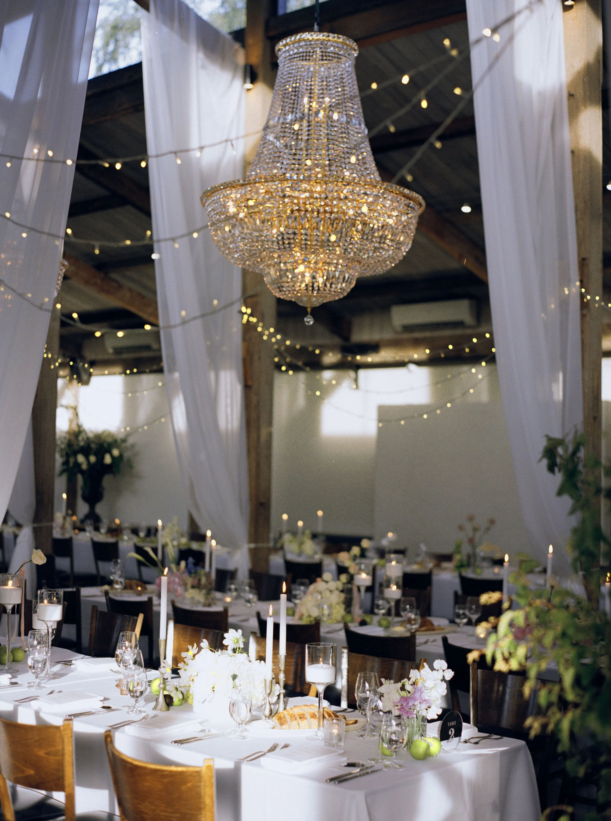 Elegant indoor wedding reception decorated with a large crystal chandelier, white drapery, string lights, and floral centerpieces on tables.