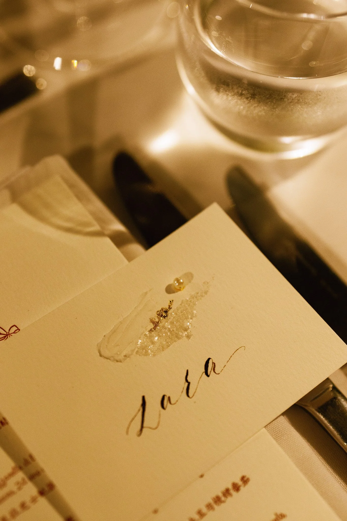 A white card with the word 'Love' written in gold cursive, decorated with small pearls and glitter. A glass of water and a necklace are visible in the background.