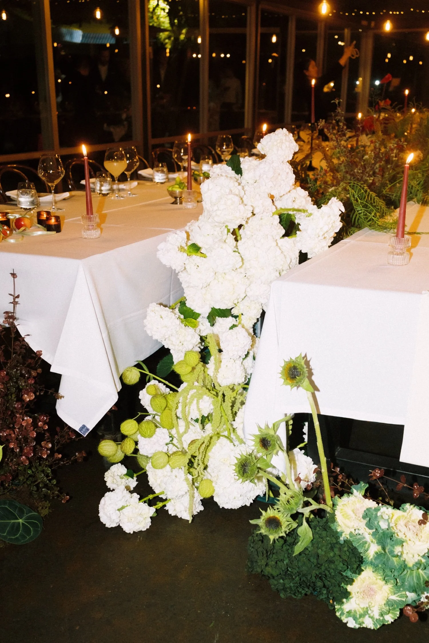 A banquet table decorated with white flowers, candles, and glasses, with a floral arrangement and a background of glass windows and people.