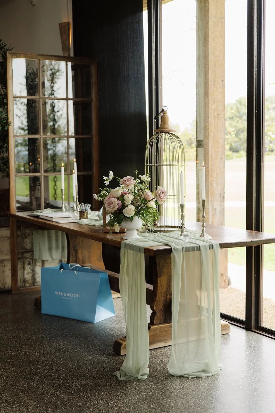 A rustic wooden table decorated with pink and white flowers, white candles, and light green drapery near large windows.