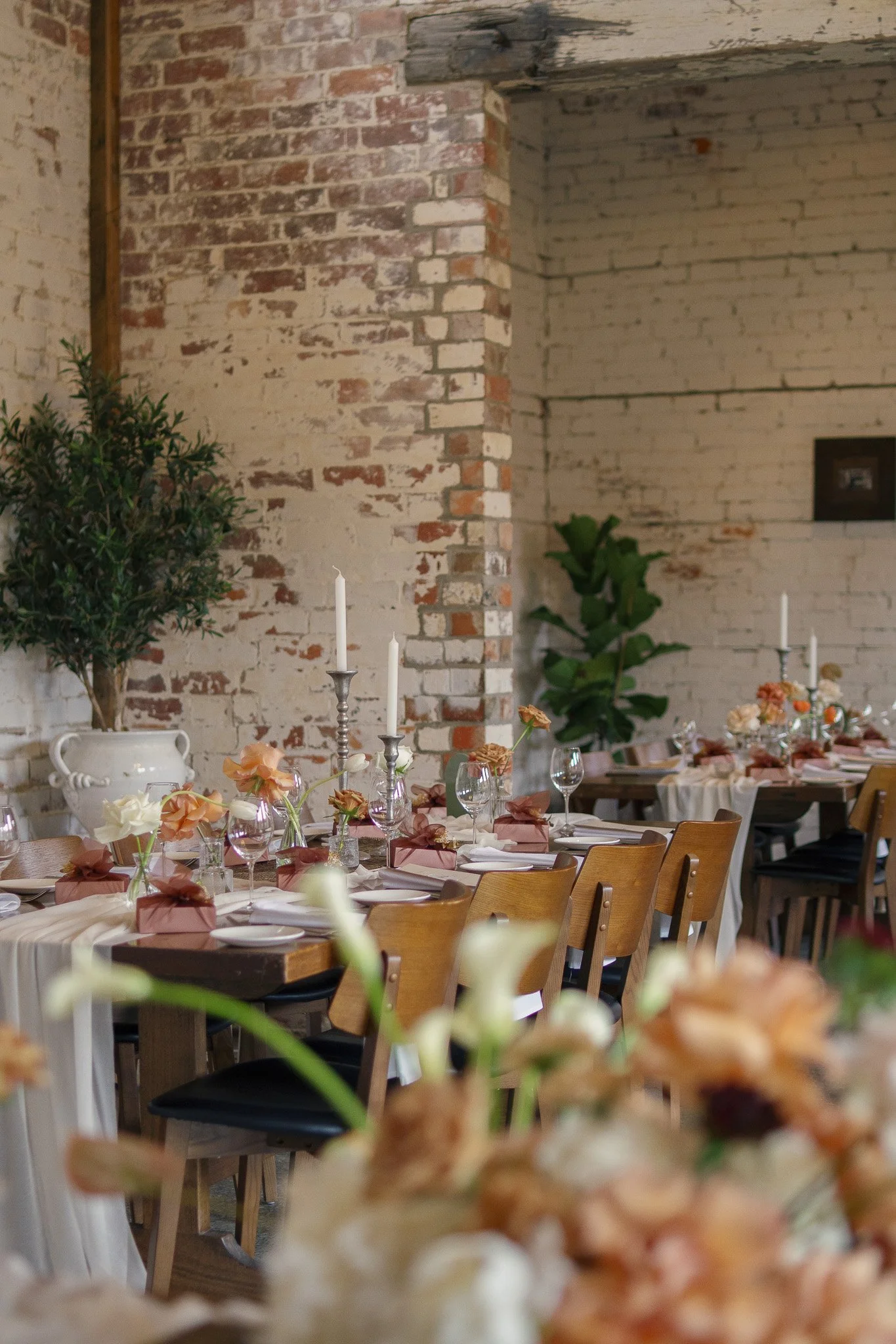 A decorated indoor dining setup with wooden chairs, white tablecloths, floral centerpieces, candles, and brick walls.