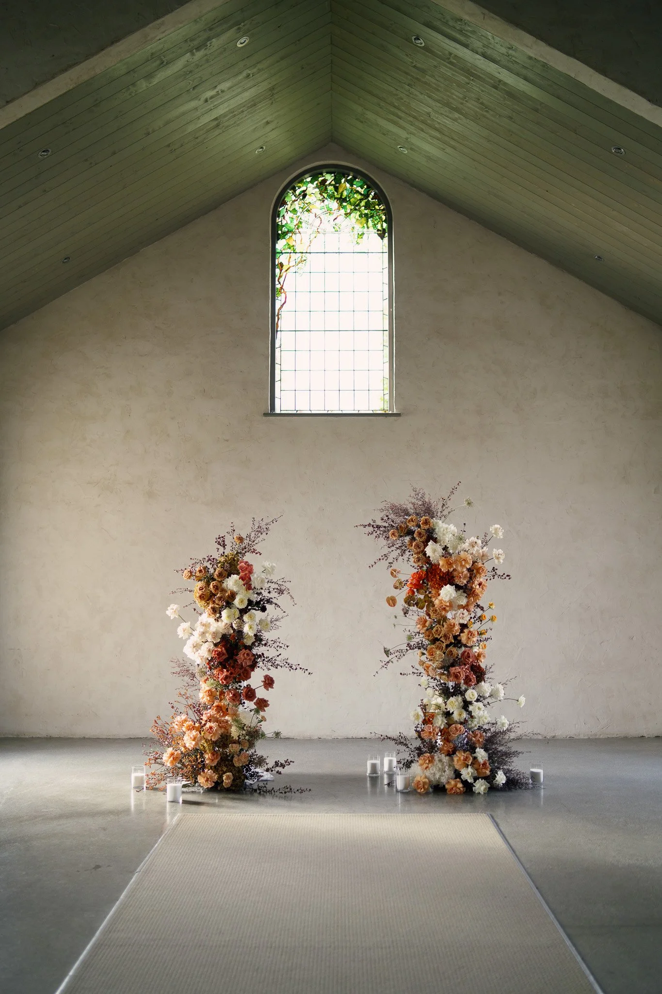 A decorated ceremony space with two large floral arrangements featuring pastel-colored roses and other flowers, placed on either side of a white aisle runner, inside a building with a high, sloped ceiling with wooden panels and a tall, arched stained