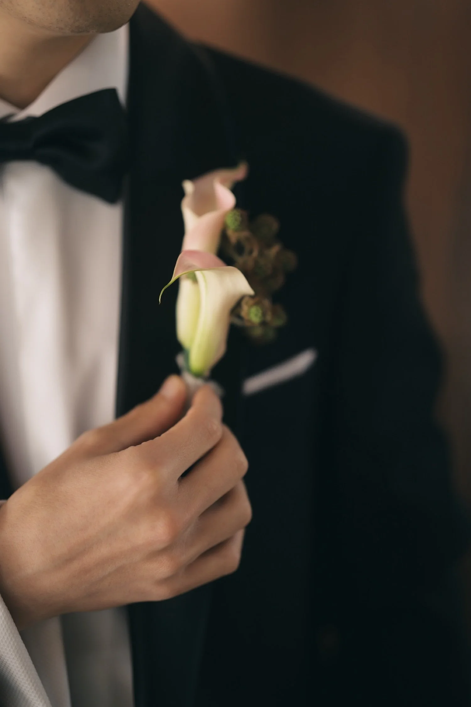 A person in a tuxedo with a white shirt, black bow tie, and boutonniere on the lapel is adjusting their attire.