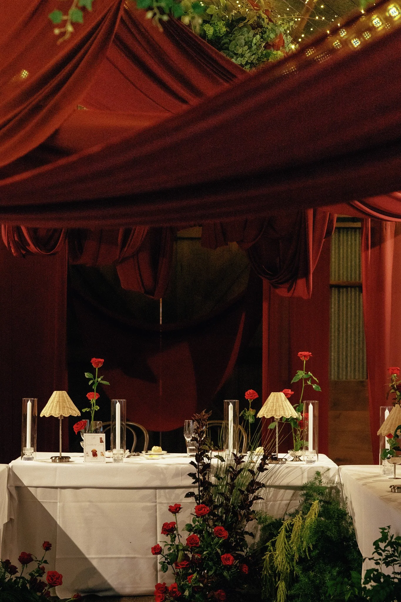 A decorated banquet table in a dimly lit room with red curtains and green plants, featuring red roses and small lamps with beige lampshades.