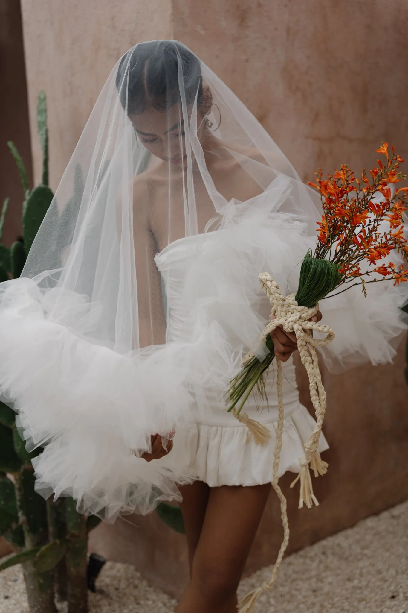 A woman dressed in a white wedding gown with a veil, holding a bouquet of orange flowers, standing indoors with a cactus and a wall in the background.