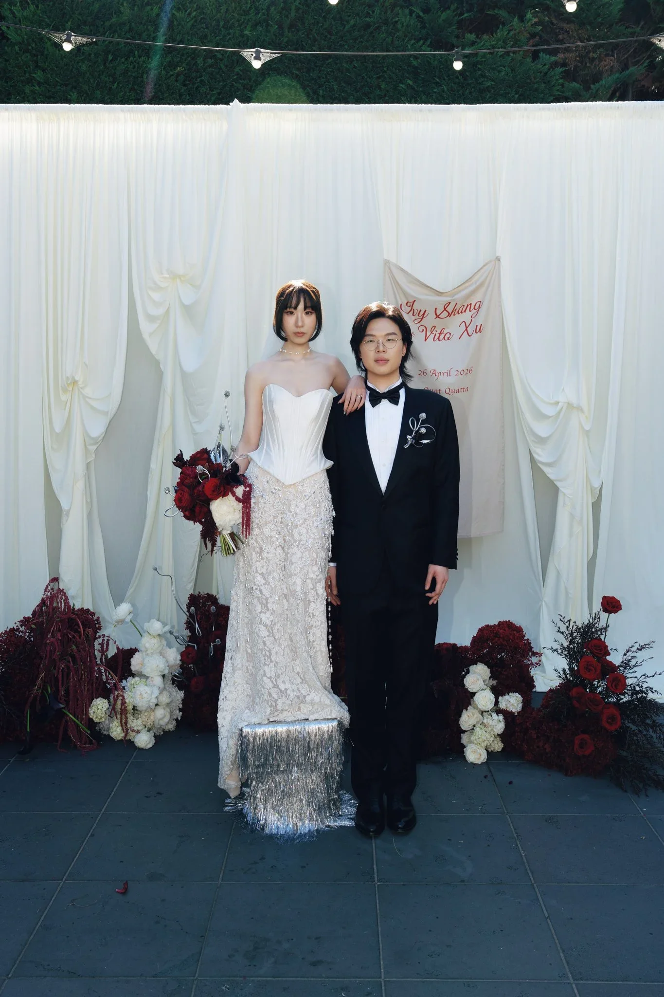 A bride and groom stand together at their wedding, with the bride in a white dress holding a bouquet, and the groom in a black tuxedo with glasses. They are surrounded by floral arrangements and are standing in front of a white backdrop with a sign t