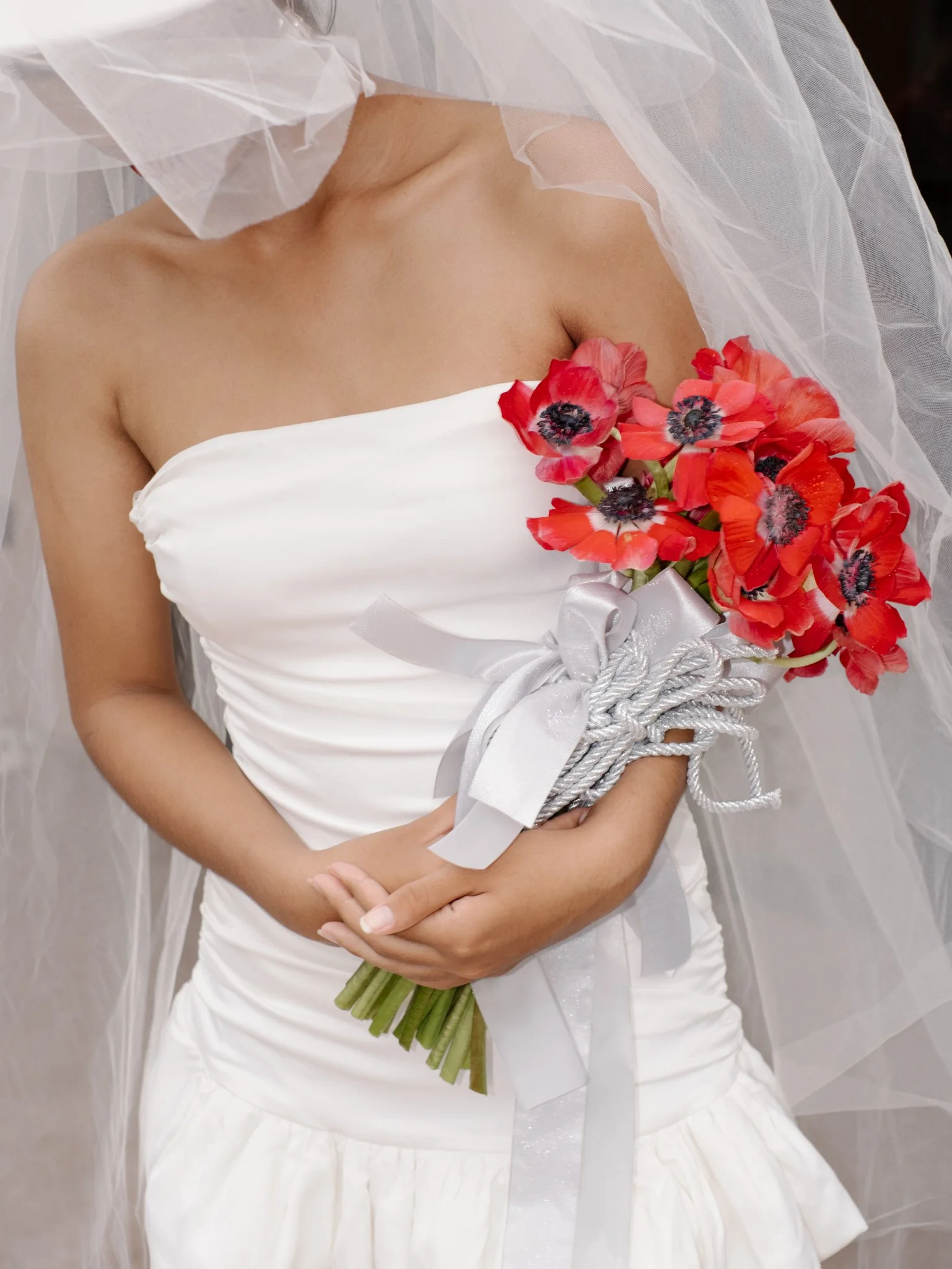 Close-up of a woman in a white strapless wedding dress holding a bouquet of red poppies, with a tulle veil partially covering her face and shoulders.