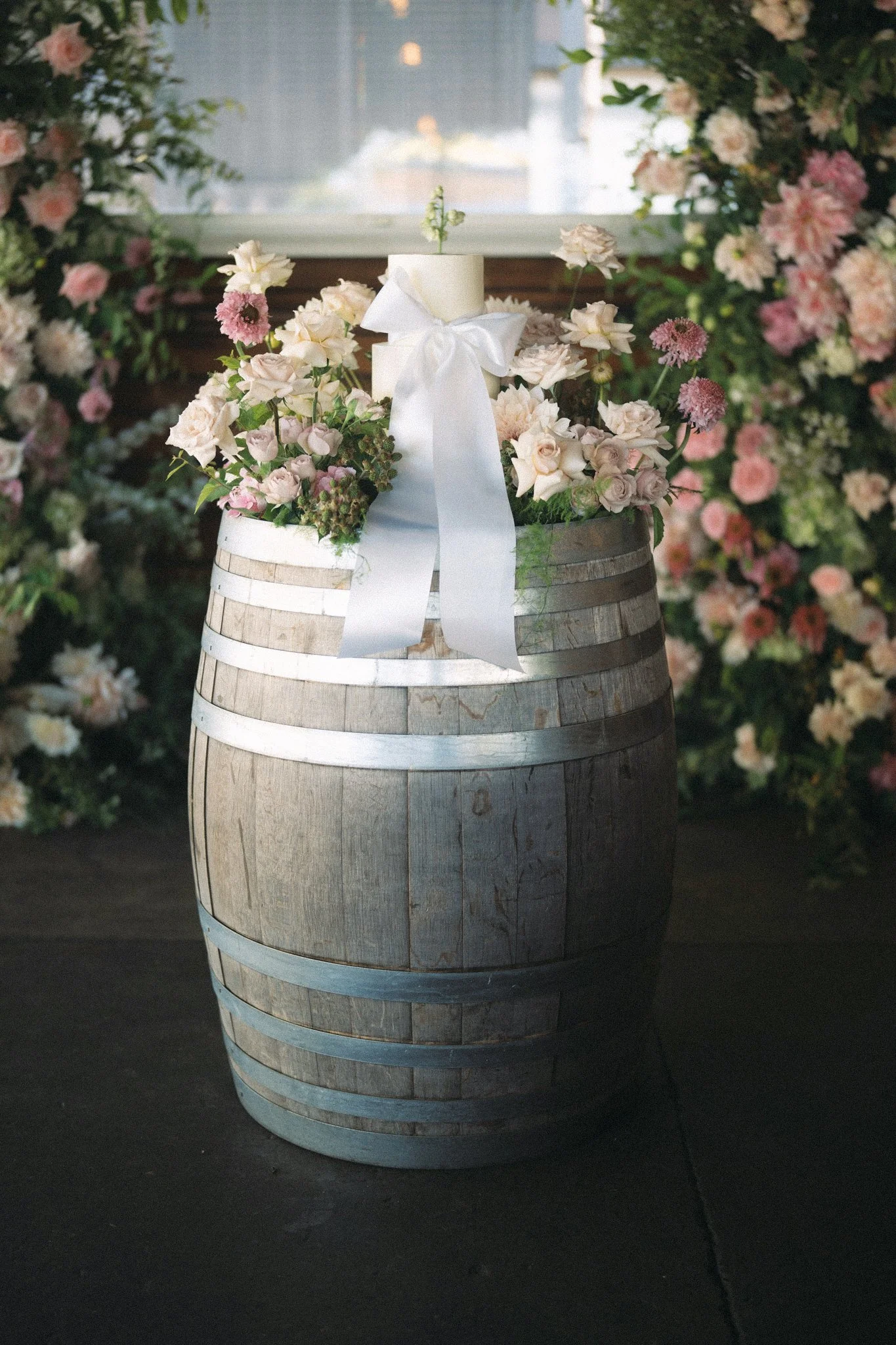 A wooden barrel with metal bands decorated with a floral arrangement of white, pink, and peach-colored flowers, topped with a white ribbon and bow. In the background, there's a window with a faint view of the outside, and the surrounding area is fill