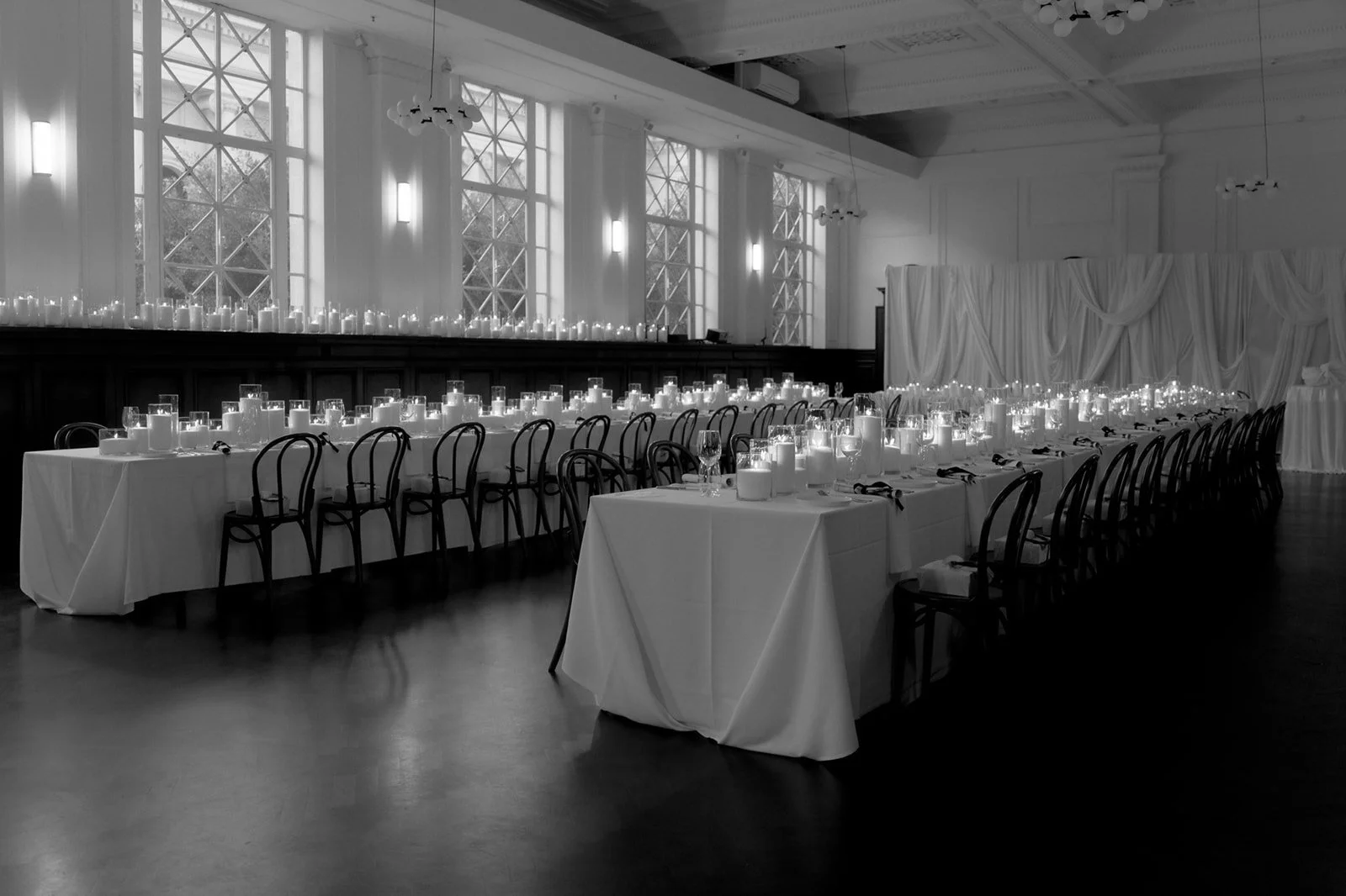Elegant banquet hall with long tables draped in white tablecloths, set with candles and glassware, high windows, and draped curtains in the background.