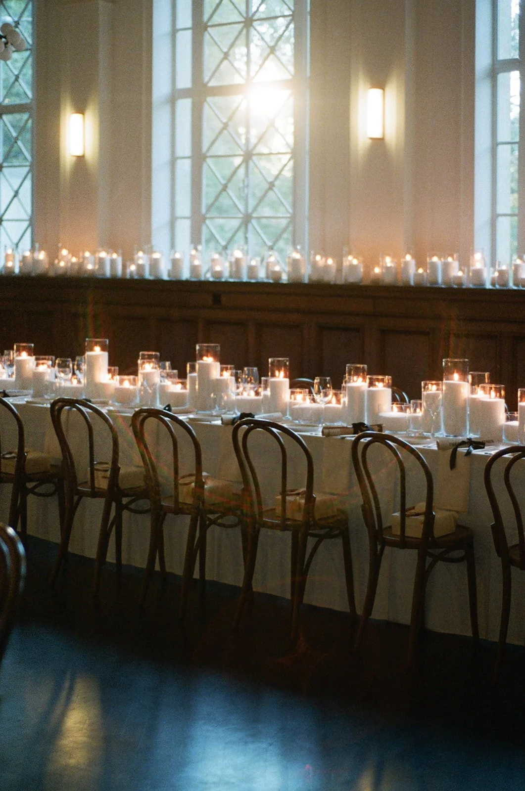 A long dining table set for a formal event with white tablecloths, lit candles in glass holders, and chairs arranged around it in a room with large windows and soft lighting.