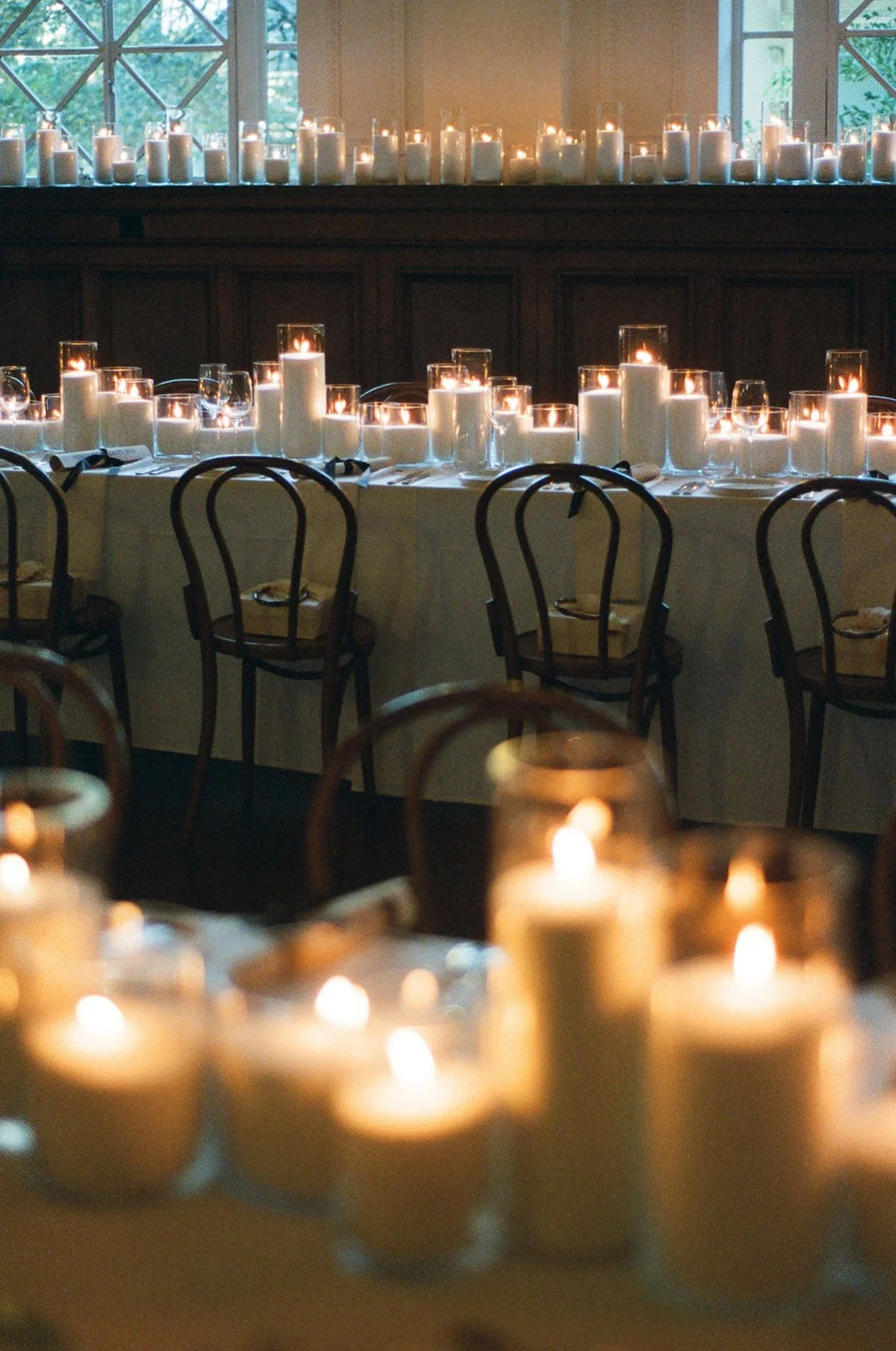 A dimly lit indoor dining area decorated with numerous white candles in glass holders, arranged on tables and a ledge, creating a warm and cozy ambiance.