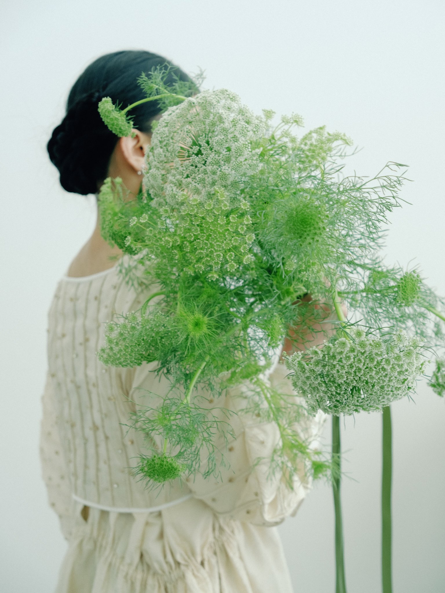 A woman with dark hair in a braid holds a large bouquet of white and green flowers, partially obscuring her face, against a plain light background.