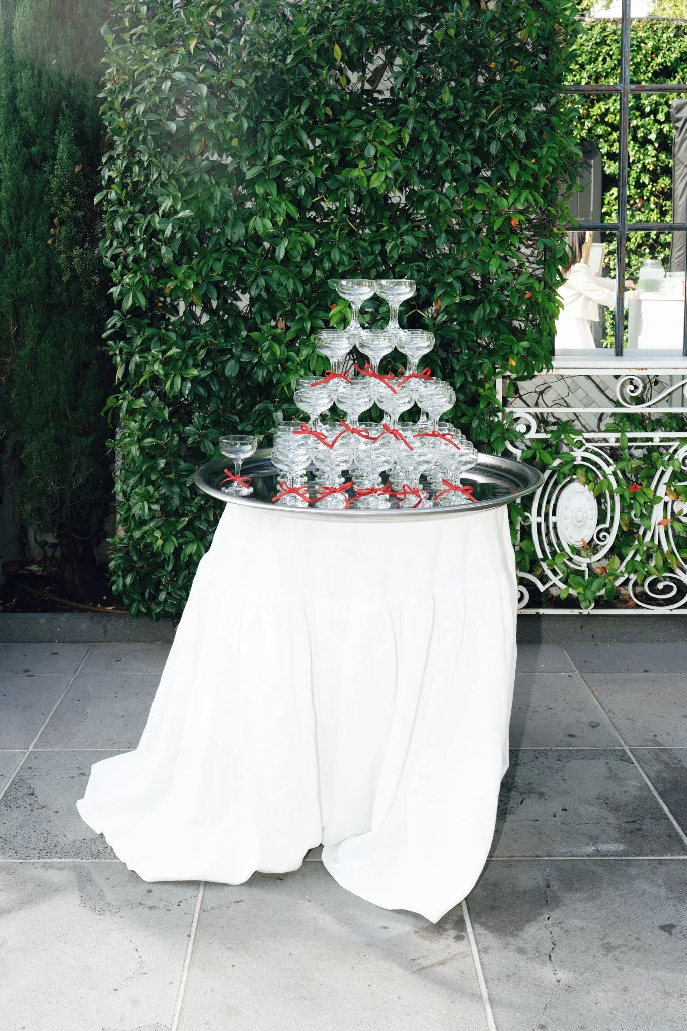 A table covered with a white cloth holding a pyramid of glass champagne coupes, some tied with red ribbons, set outdoors against a green hedge and a window with a white railing.