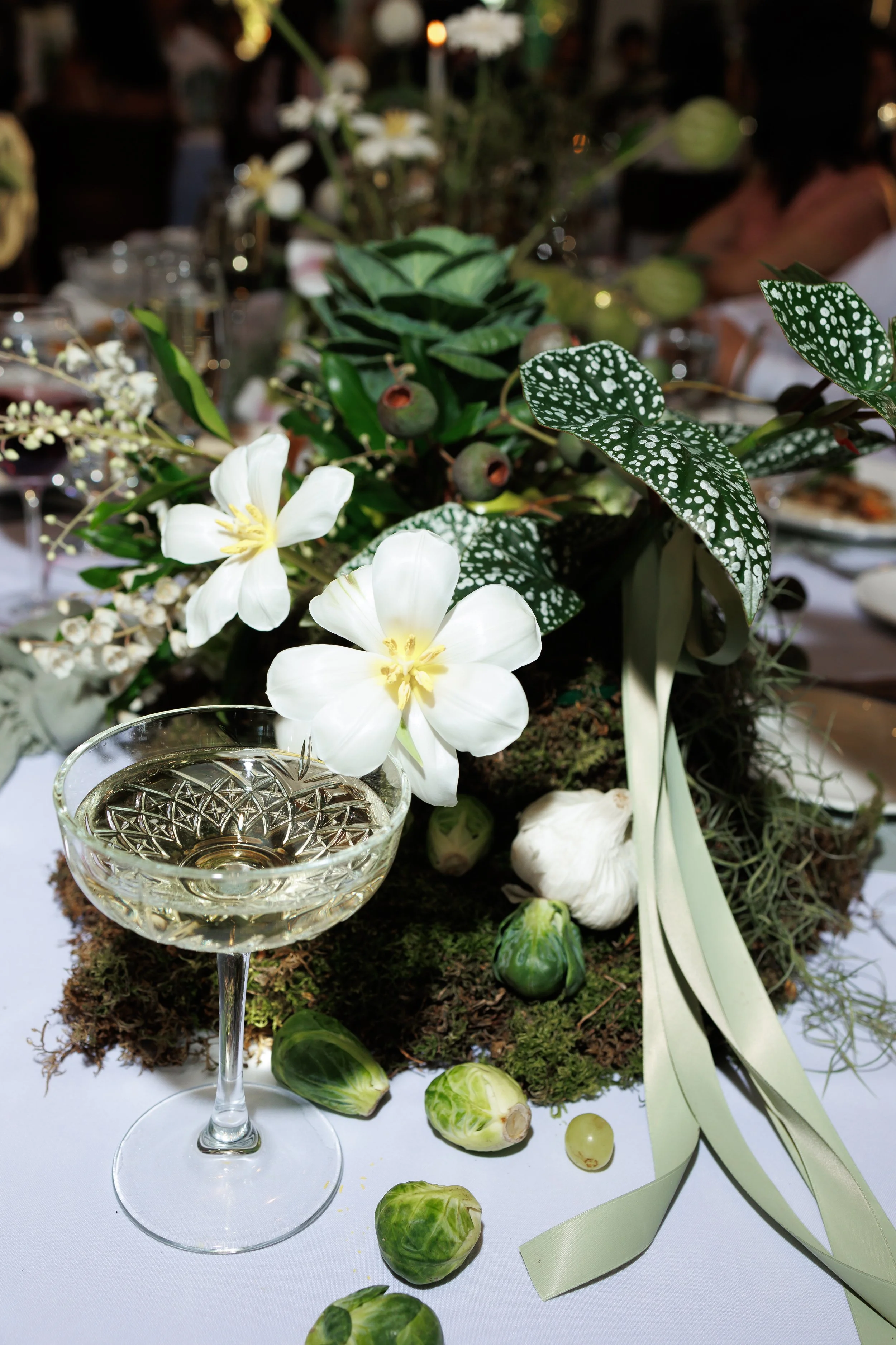 A table centerpiece with white flowers, green leaves, and small green fruits, set on a white tablecloth with scattered vegetables and a glass of white wine in the foreground.