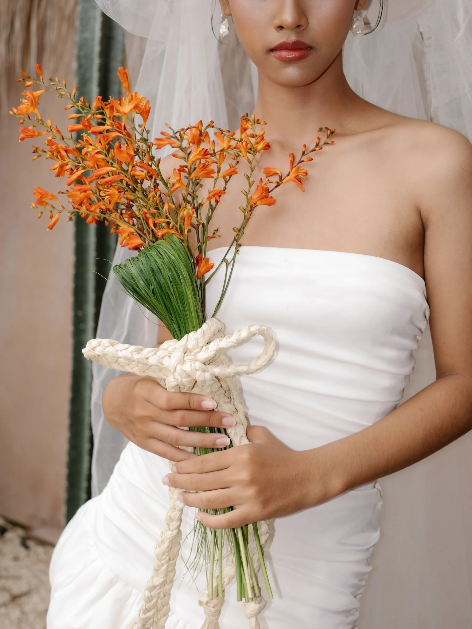 A woman dressed in a white strapless dress holding a bouquet of orange flowers with green leaves, wrapped in a white woven band.