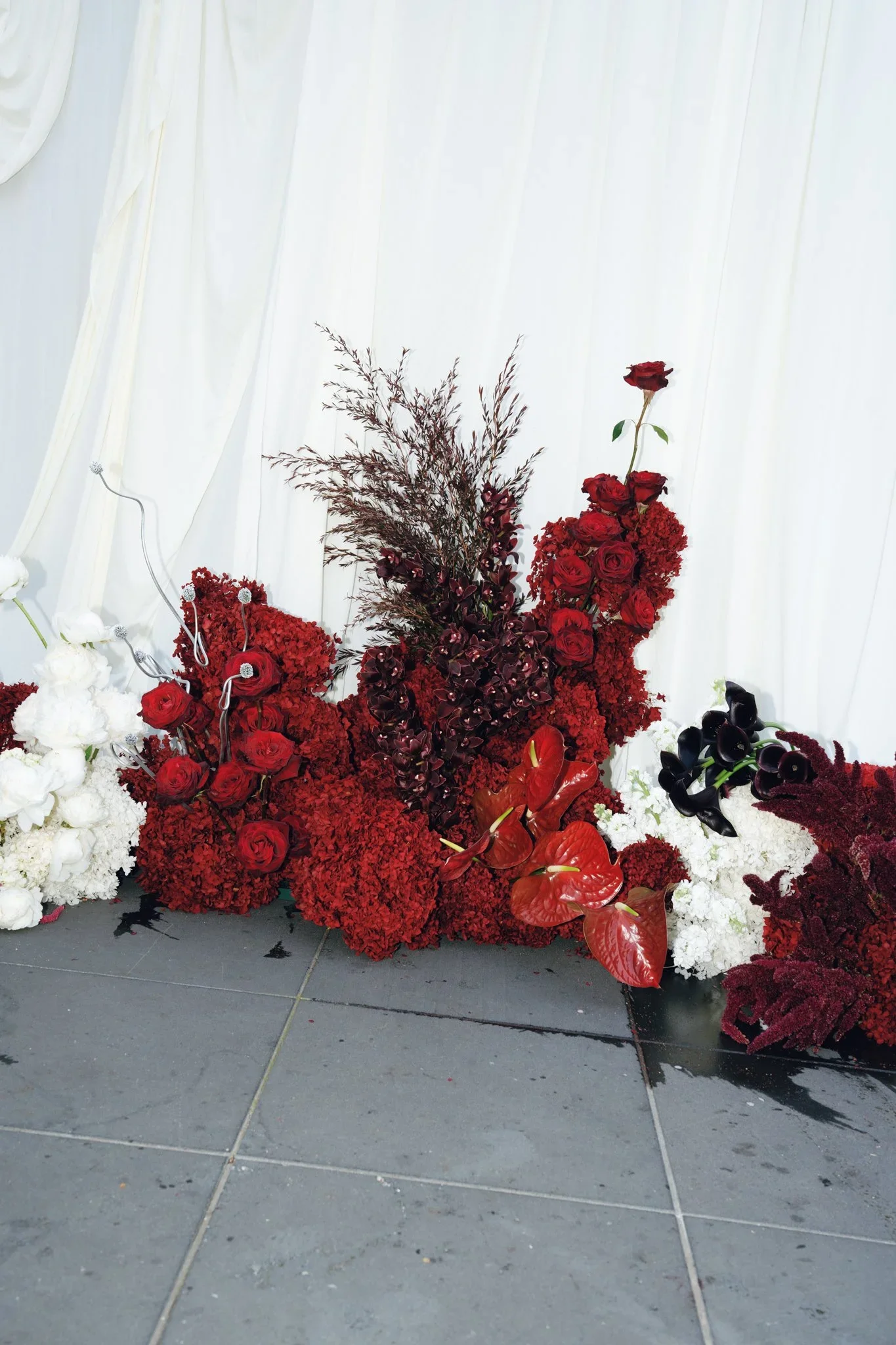 A floral arrangement featuring red and white flowers, including roses and anthuriums, displayed against white draped fabric.