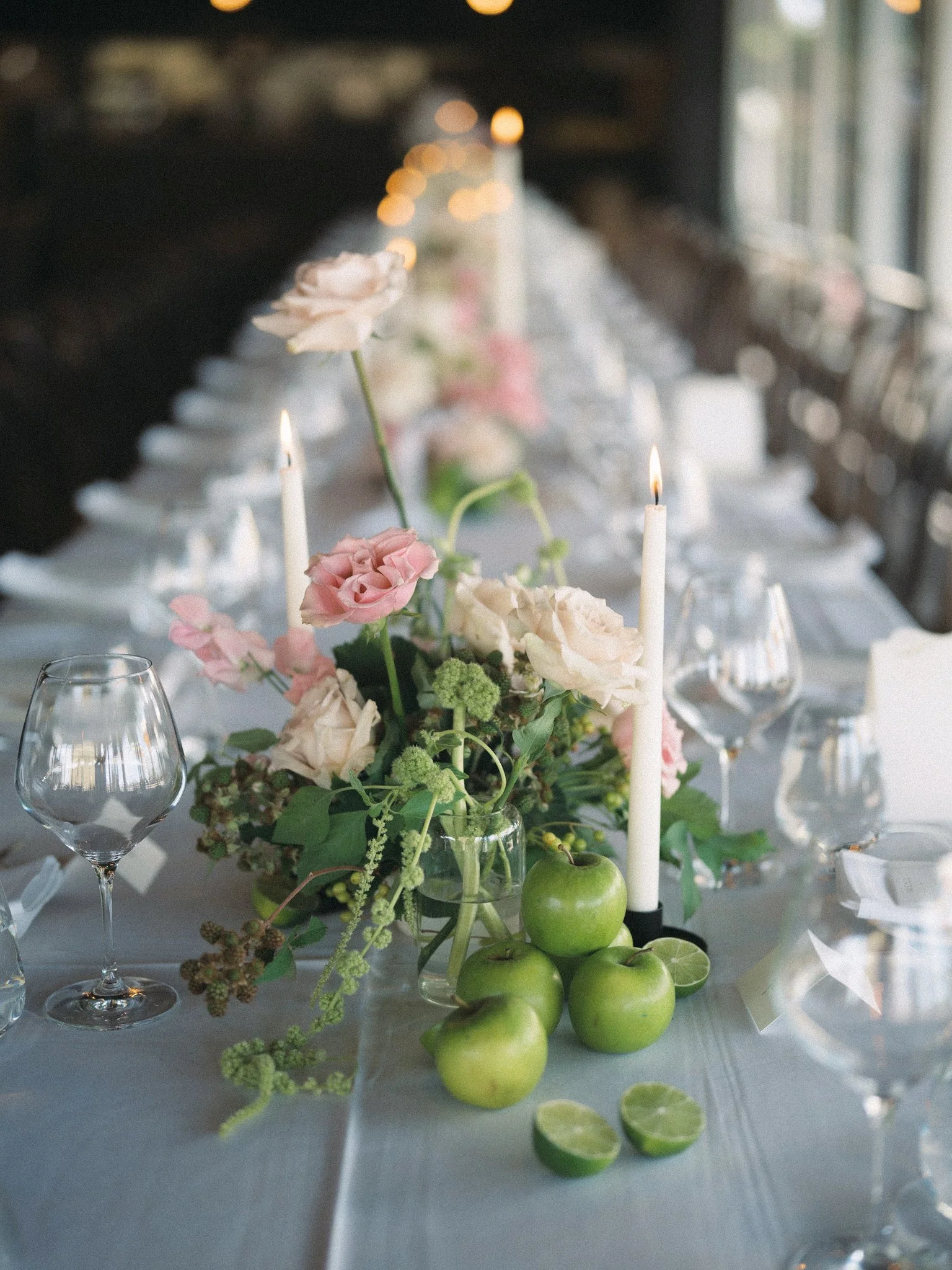 A table decorated with a floral arrangement of pink and cream flowers, green apples, and greenery, illuminated by candles with burning wicks, set against a blurred background of string lights and windows.