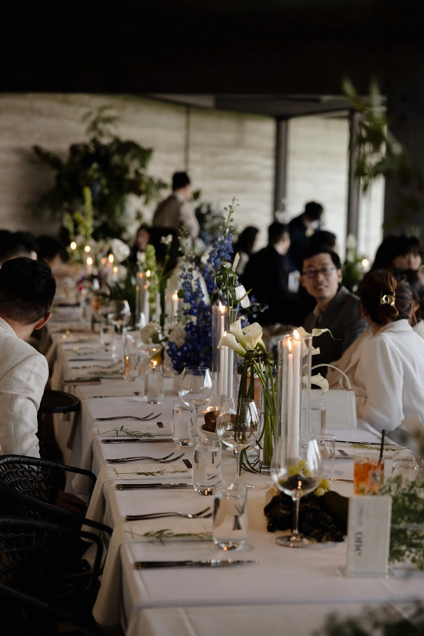 A long banquet table set for a formal event with white tablecloths, tall white candles, and floral centerpieces featuring white and blue flowers, surrounded by guests in formal attire inside a modern venue with large windows and greenery.