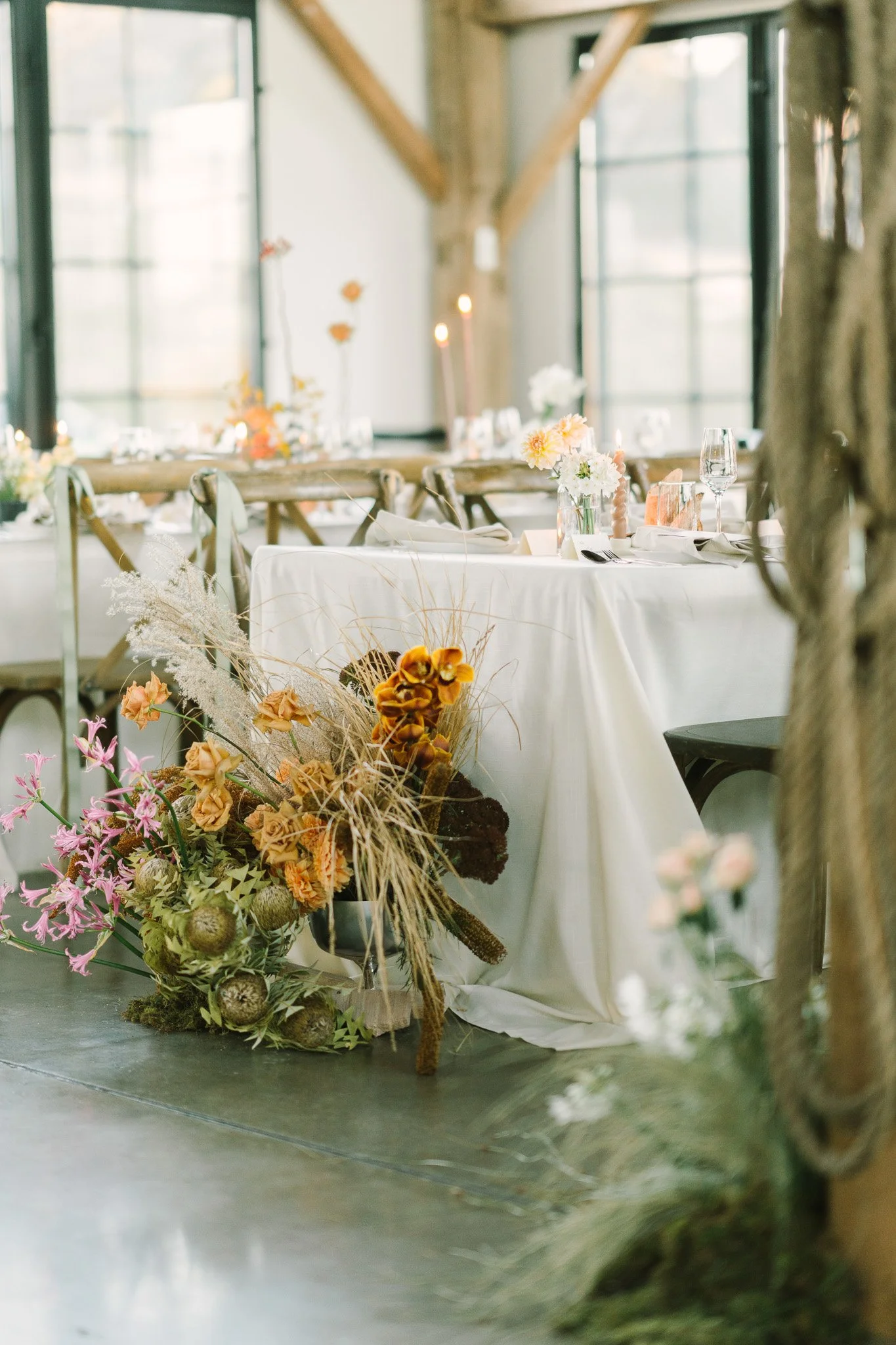 A decorated indoor event space with a long table adorned with a floral centerpiece in the foreground. The background features large windows letting in natural light, and the table is set with glassware, candles, and tableware.