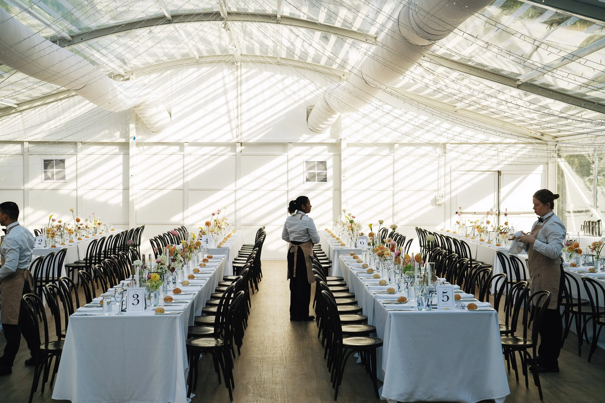 A banquet hall with long tables covered in white tablecloths, decorated with floral centerpieces, set with utensils and bread rolls. There are black chairs along the tables, and waitstaff are preparing the venue. The ceiling has a glass roof with sun