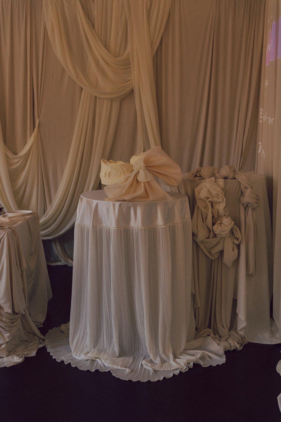 Elegant beige draped curtains with a round table covered in matching beige fabric, topped with a bow-shaped decoration and surrounded by other draped fabric-covered items in a luxurious setting.