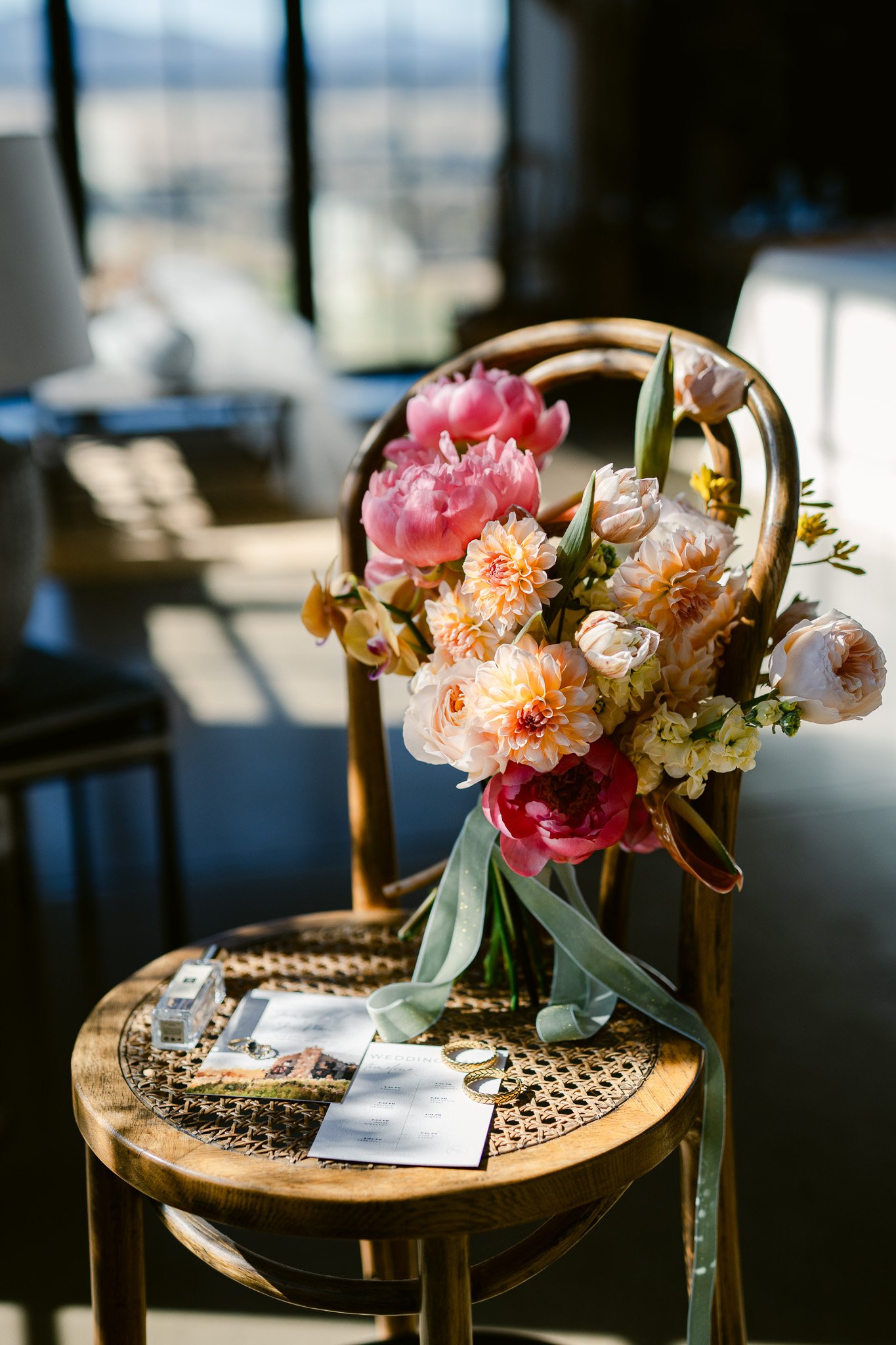 A wooden chair with a bouquet of pink and peach flowers hanging on the backrest, placed beside a round table with wedding magazines, jewelry, and a wedding planning booklet inside a room with sunlight and window shadows.