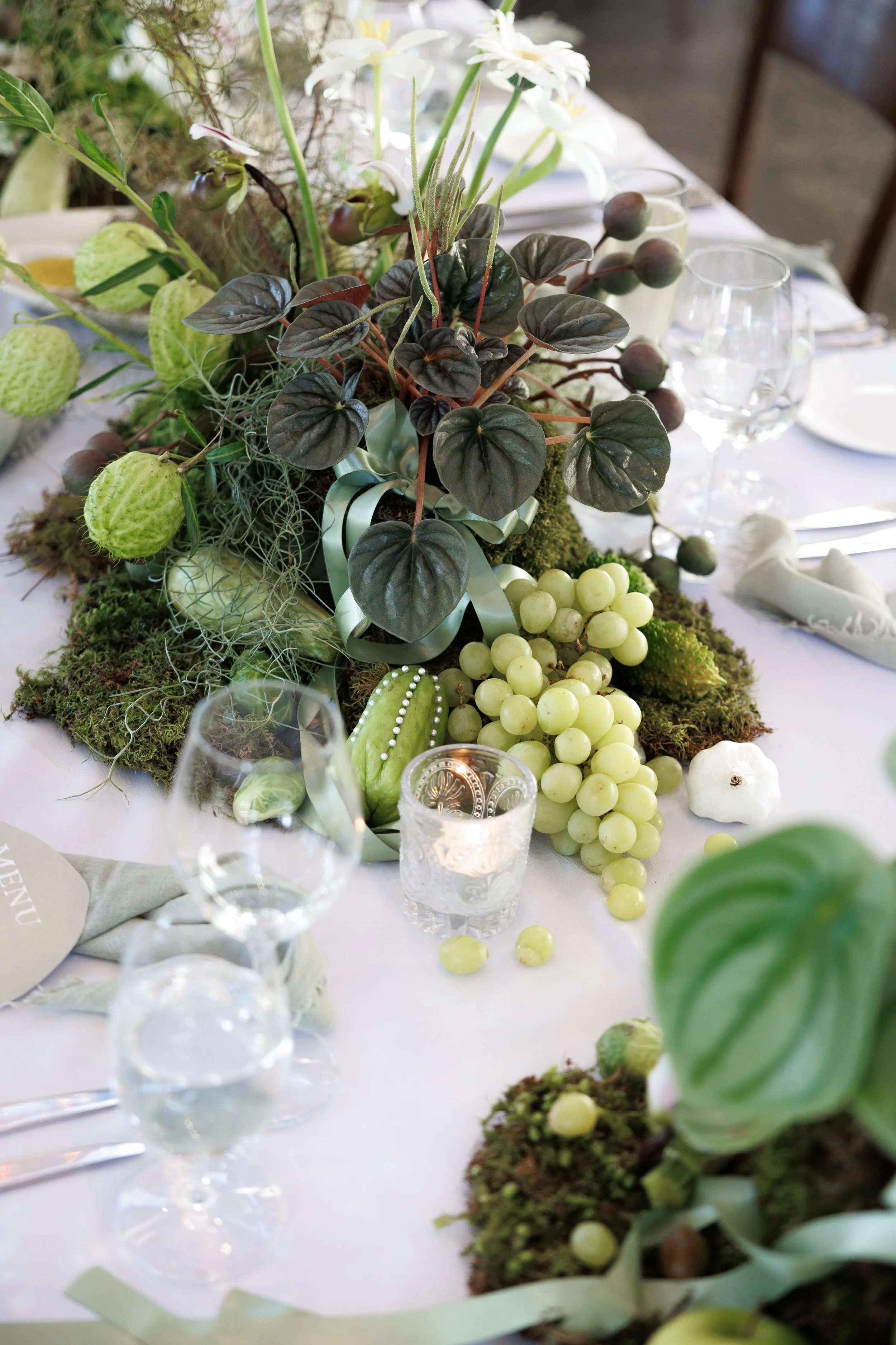 Decorative centerpiece with green moss, white grapes, dark green and purple leaves, and white flowers on a table set for a meal.