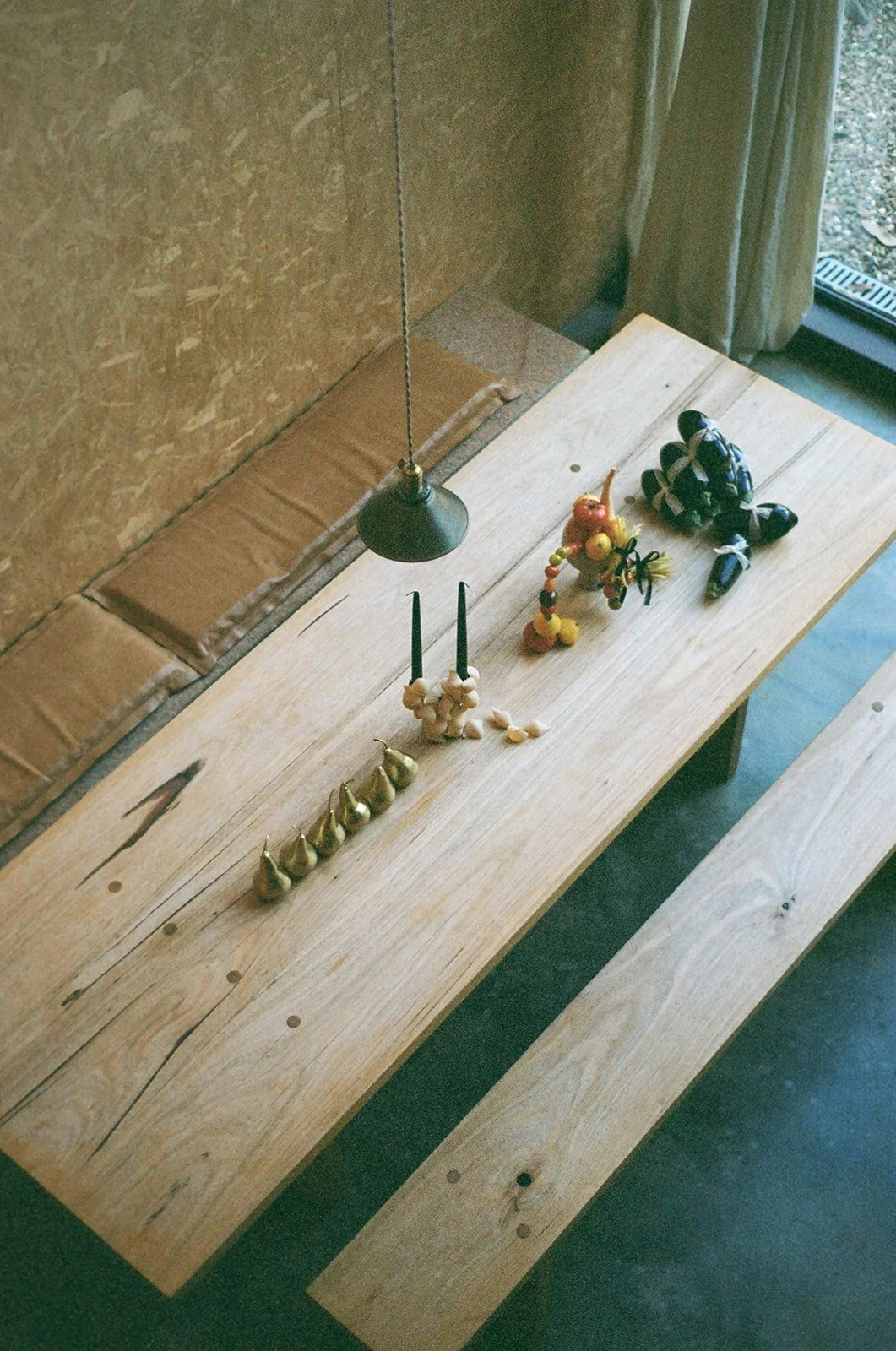 Wooden table with decorative objects, including a black hanging lamp, golden feather-shaped objects, a string of beads, a small colorful felt figure, and a black and blue rugby ball, next to a window with a curtain.