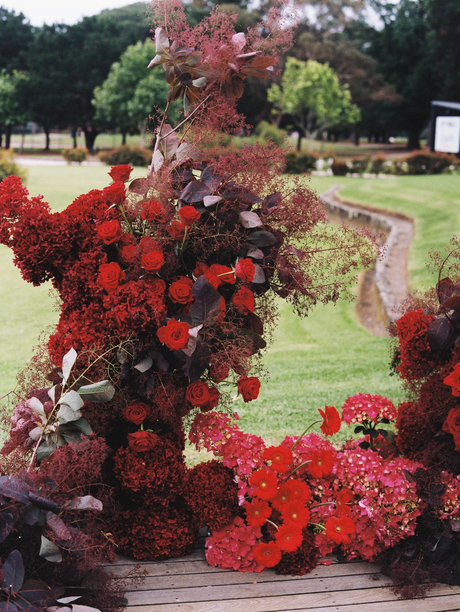 Arrangement of red roses, hydrangeas, and other red flowers in a garden setting on a wooden surface, with a grassy lawn and trees in the background.