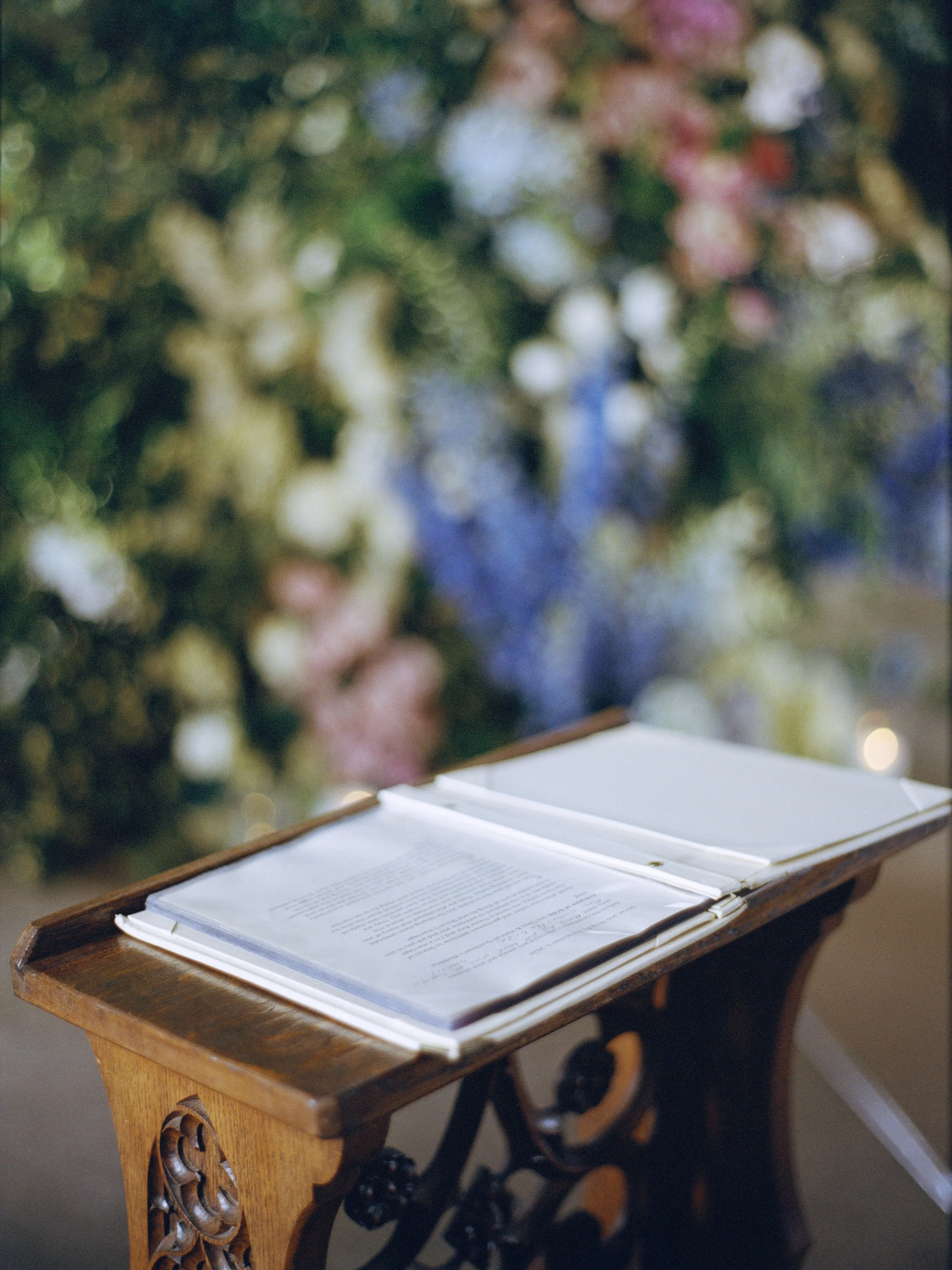 A wooden table with an open book resting on it, with a blurred background of colorful flowers and greenery.