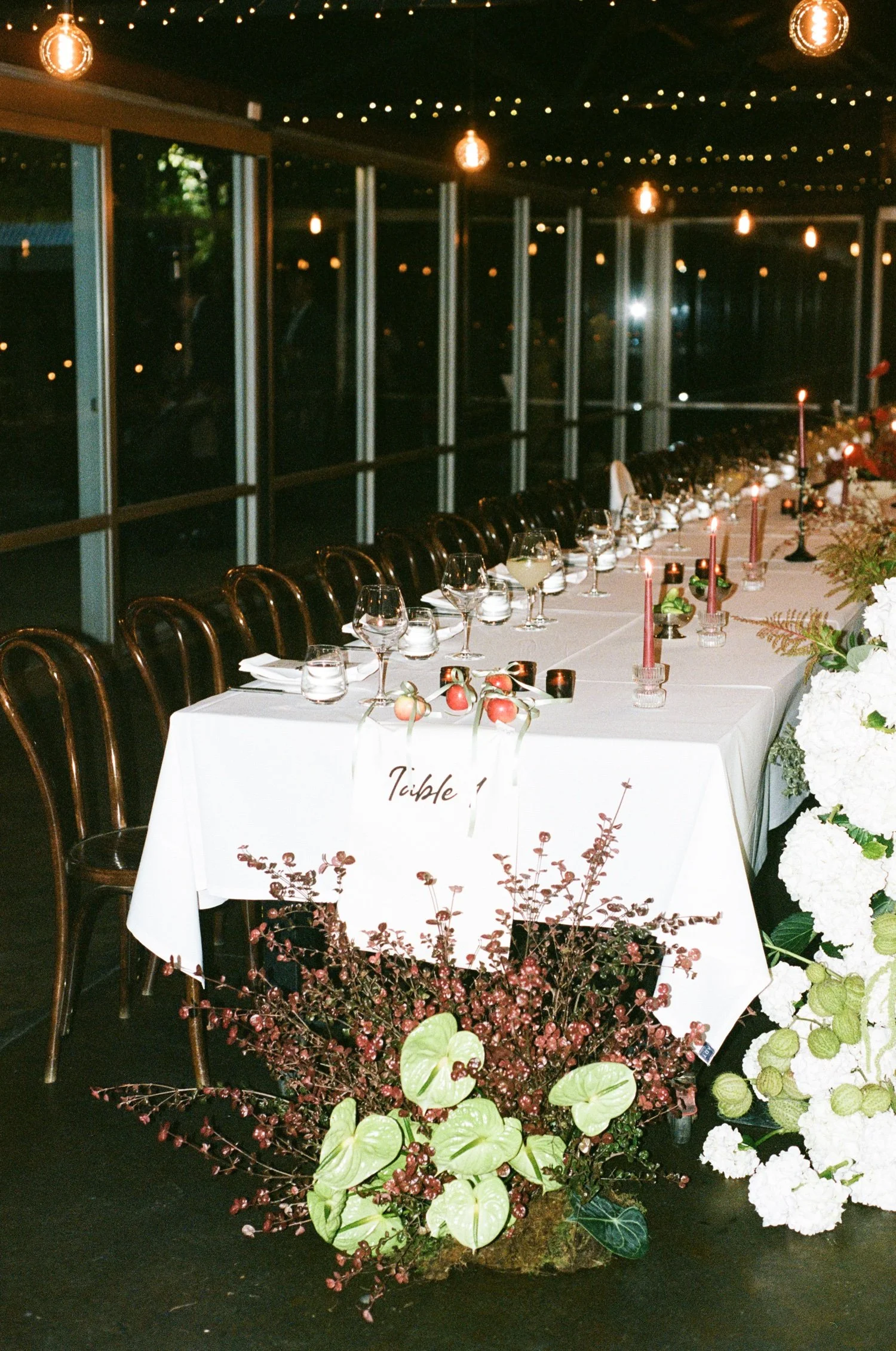 A long dining table decorated for an event, with wine glasses, candles, flowers, and table settings. Behind the table are large glass windows reflecting hanging lights and string lights in a dark room.
