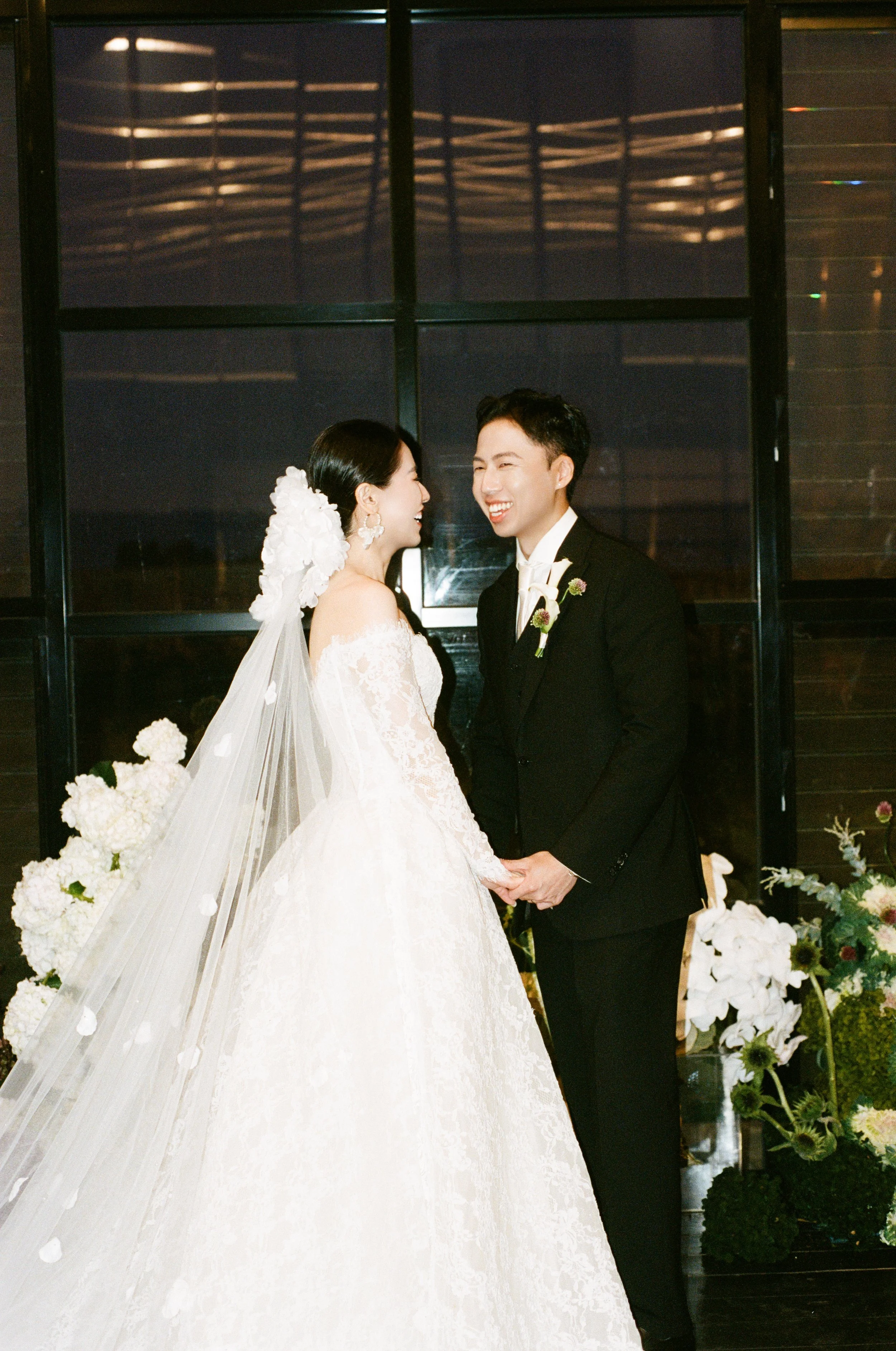 A bride and groom are holding hands and looking at each other, smiling during their wedding ceremony. The bride is wearing a white lace wedding dress with long sleeves and a floral headpiece with a veil. The groom is dressed in a black tuxedo with a 