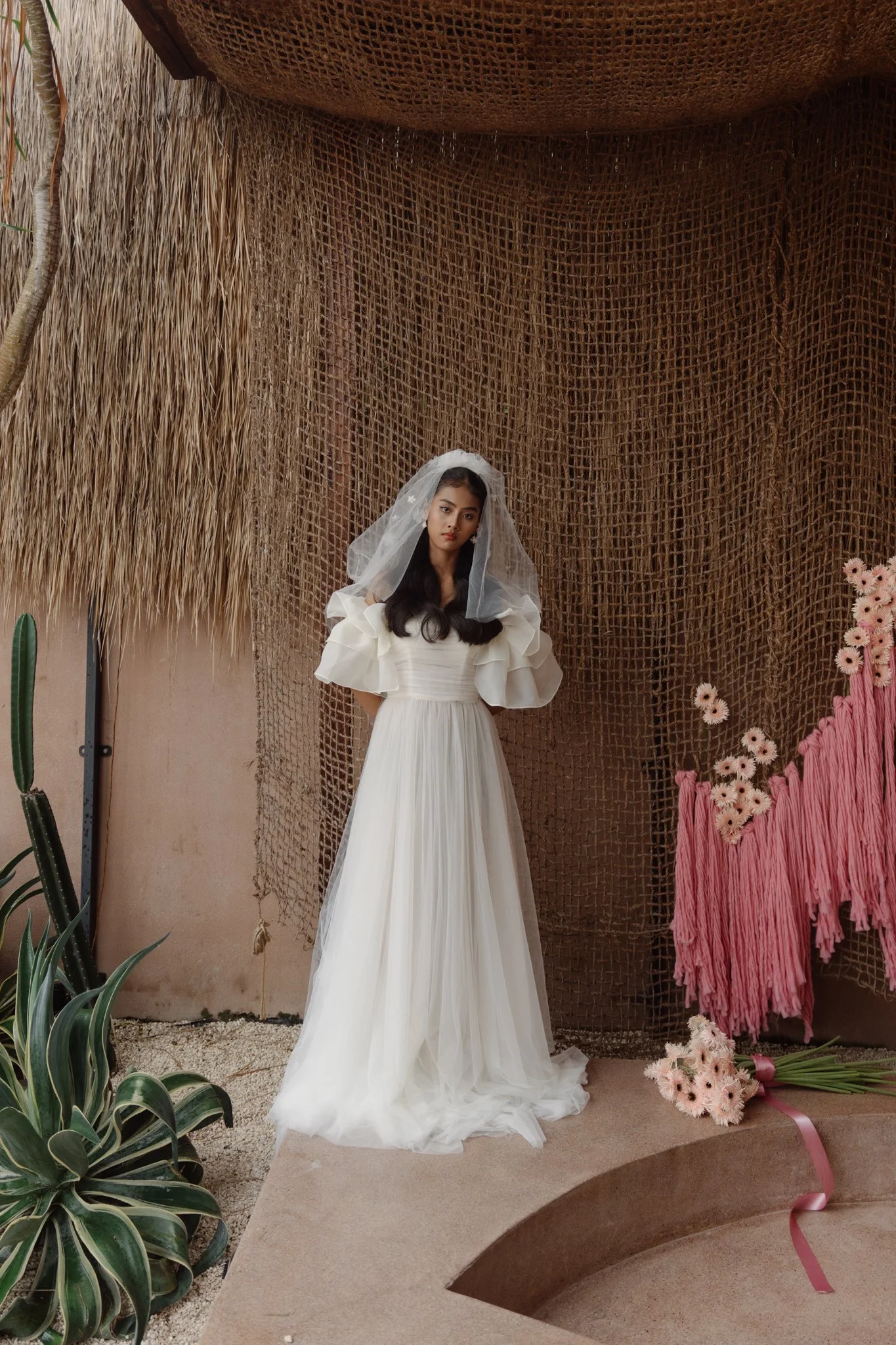 A woman dressed in a long white gown and veil standing in a decorative setting with plants, flowers, and textured brown backdrop.