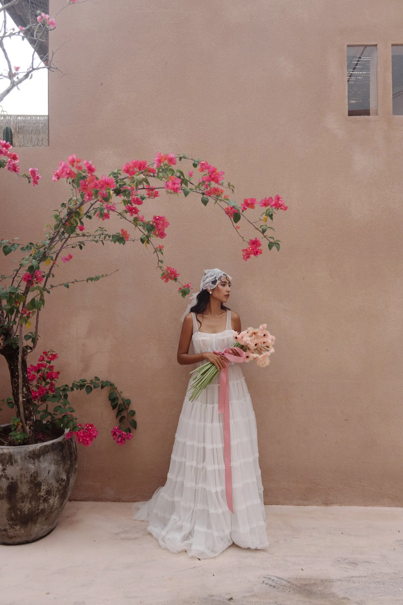 A woman in a white dress and lace headpiece holding a bouquet of pink flowers, standing against a beige wall with a large potted plant with pink flowers on her left.