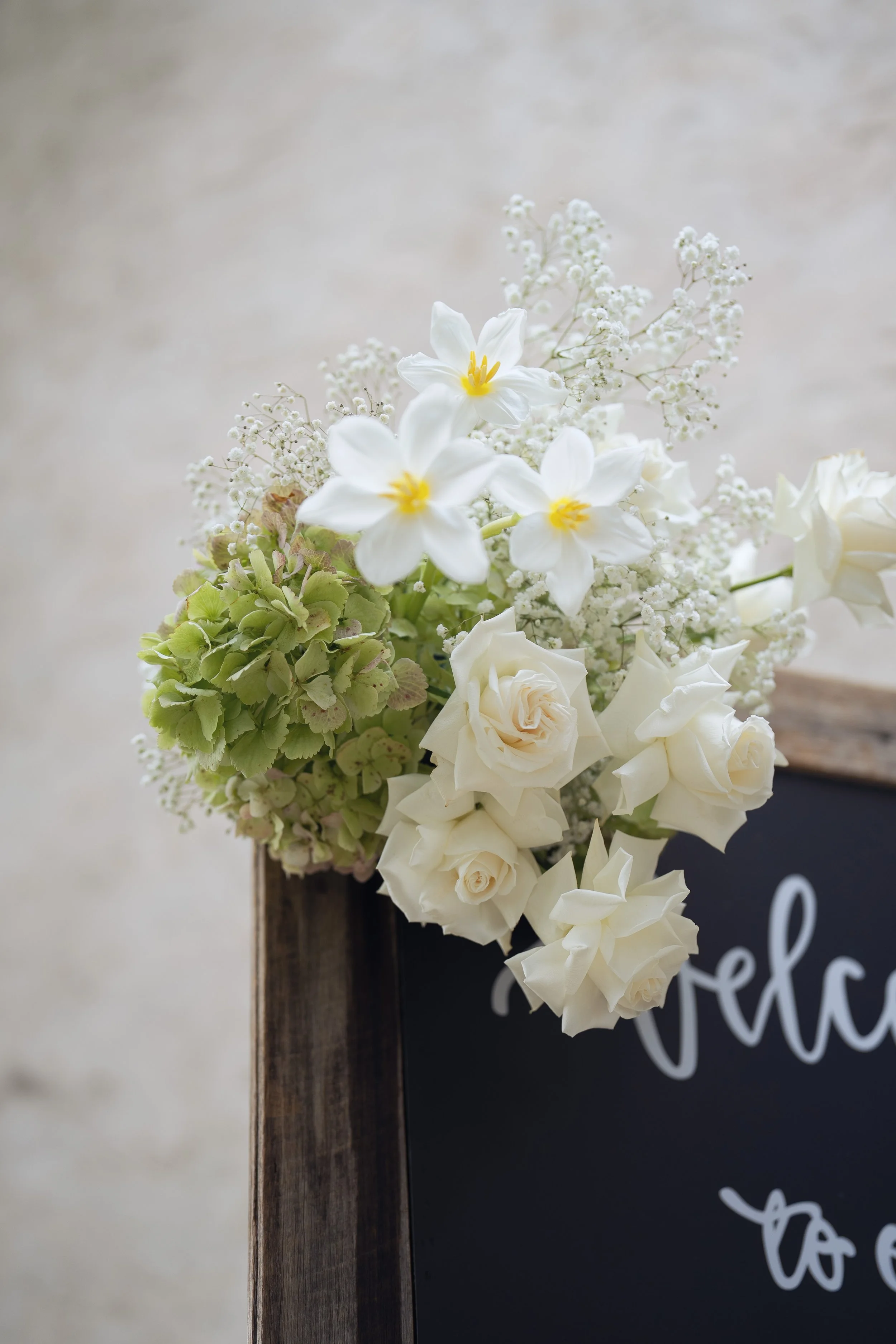 A bouquet of white flowers, including roses, daisies, and baby's breath, placed next to a wooden stand and a black sign with white lettering.