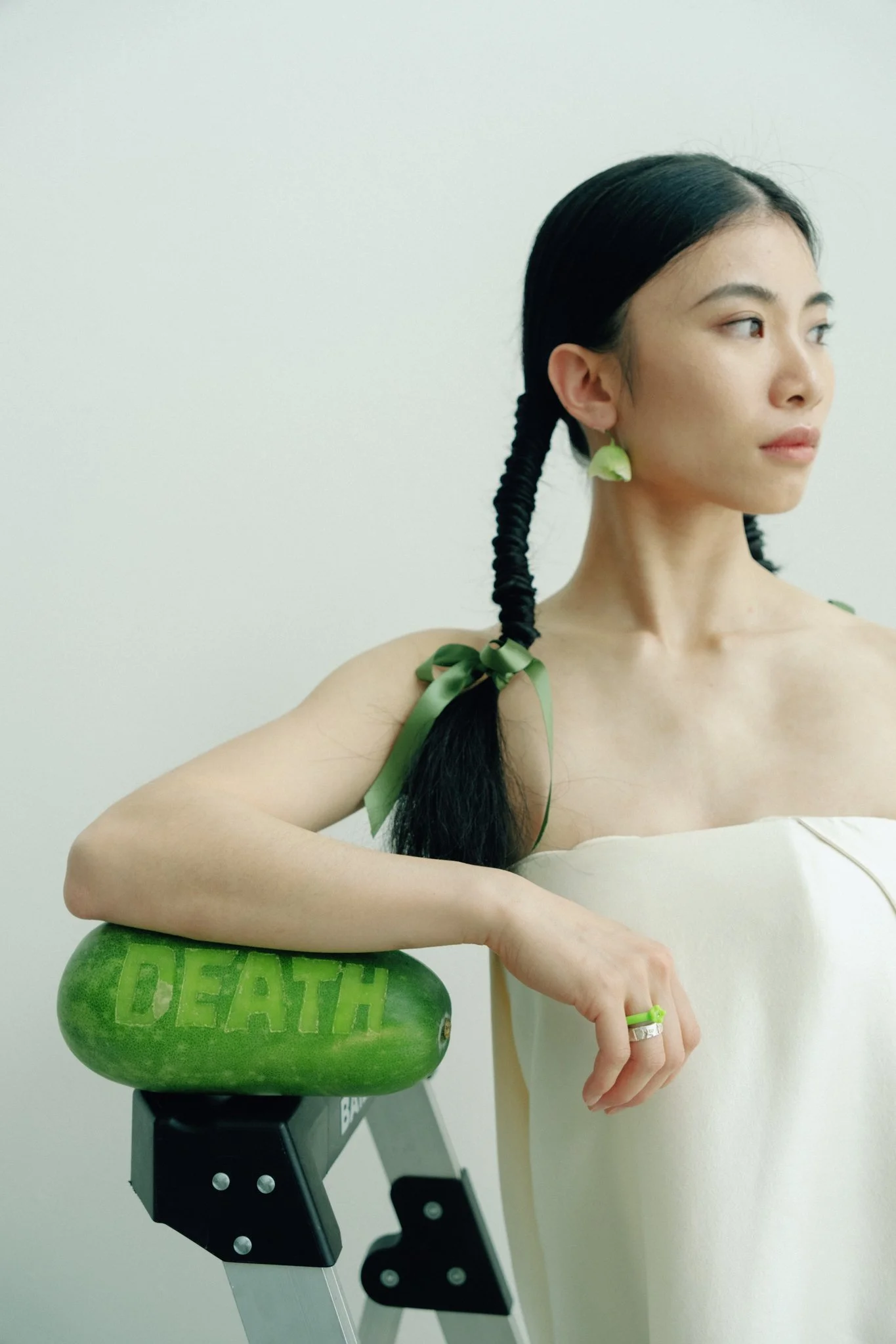 A young woman with dark hair styled in braids with green ribbons, wearing earrings made of leafy material and a ring, is sitting beside a watermelon with the word 'DEATH' carved into it, on a black bicycle or portable stand, against a plain white wal