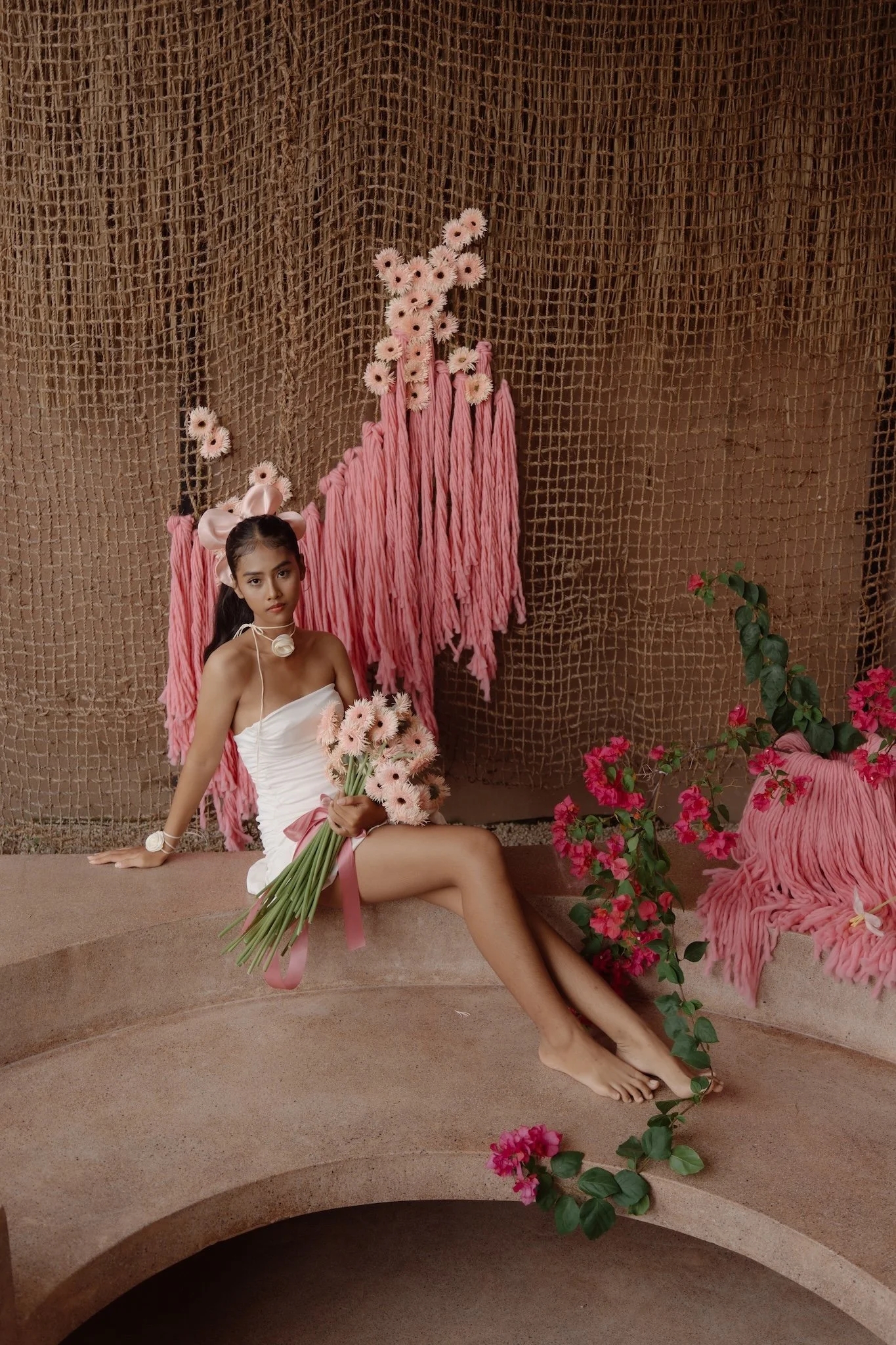 A young woman with long dark hair sitting on a curved stone ledge, holding a bouquet of pink flowers, dressed in a strapless white dress with pink ribbons, surrounded by pink and white flowers and a textured brown backdrop.