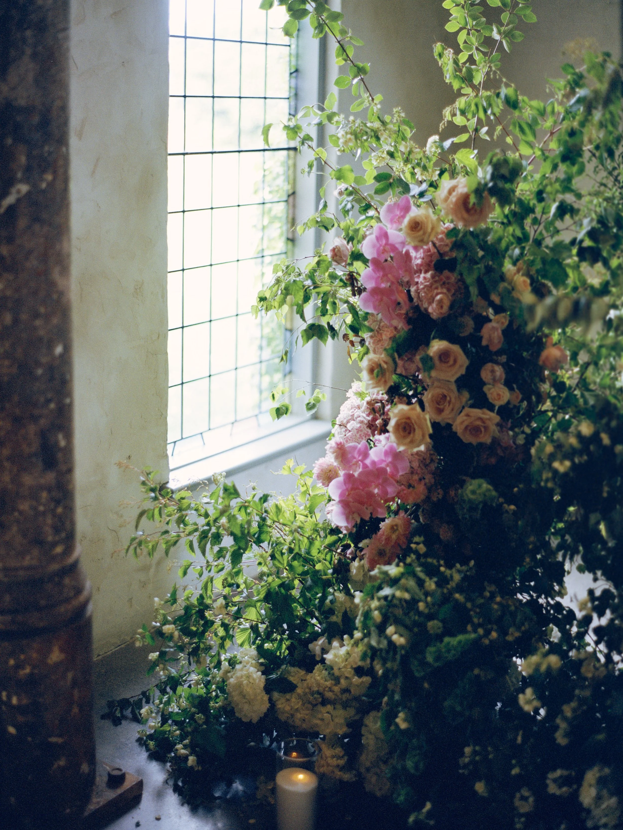 A floral arrangement with pink, cream, and white flowers beside a window with metal grid bars, lit by natural light with a candle on a dark surface in the foreground.