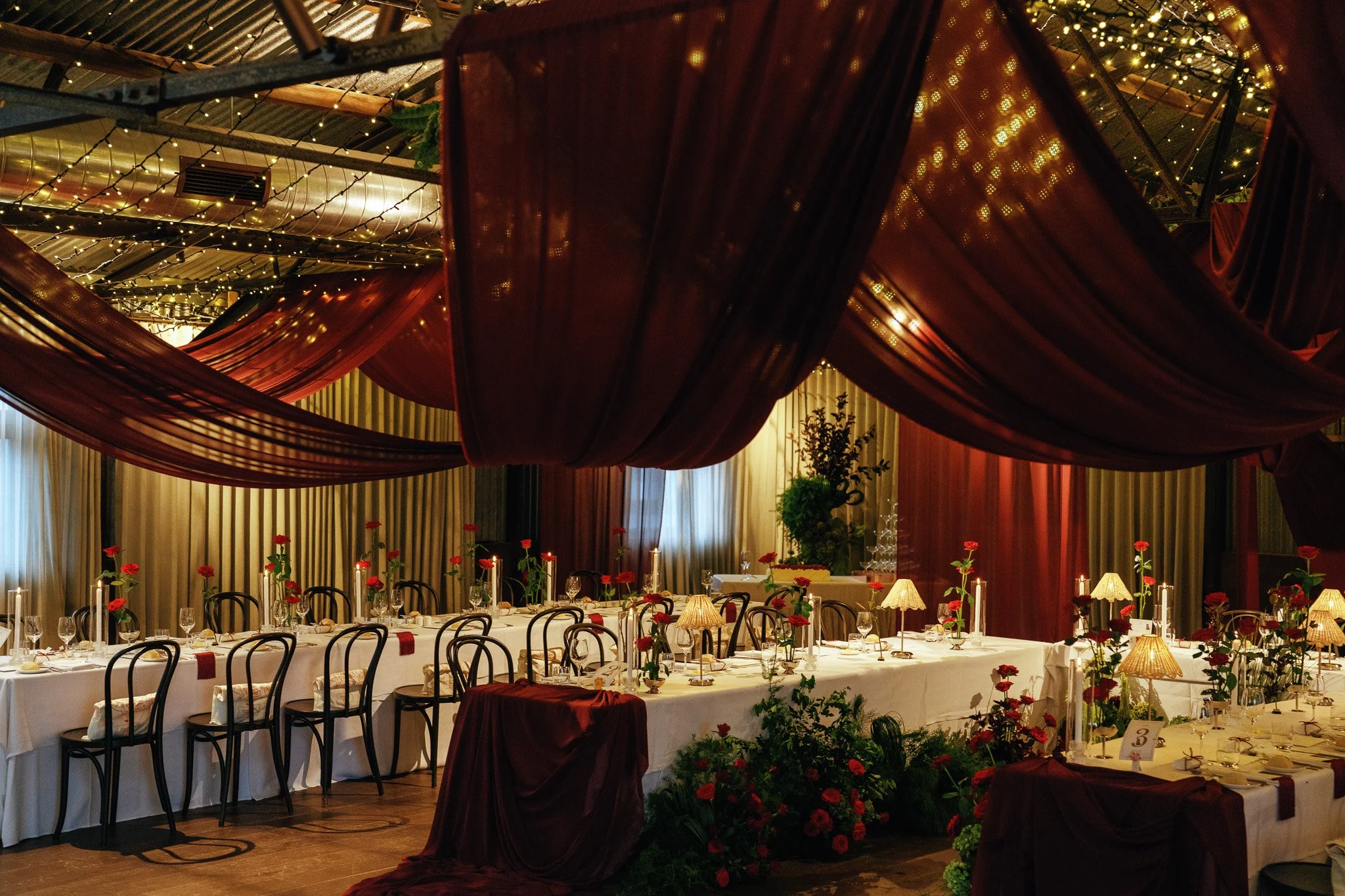 A beautifully decorated banquet hall with round tables covered with white tablecloths, adorned with red roses, candles, and small lamps. The ceiling is decorated with dark red drapes and string lights, creating a warm ambiance. The background feature