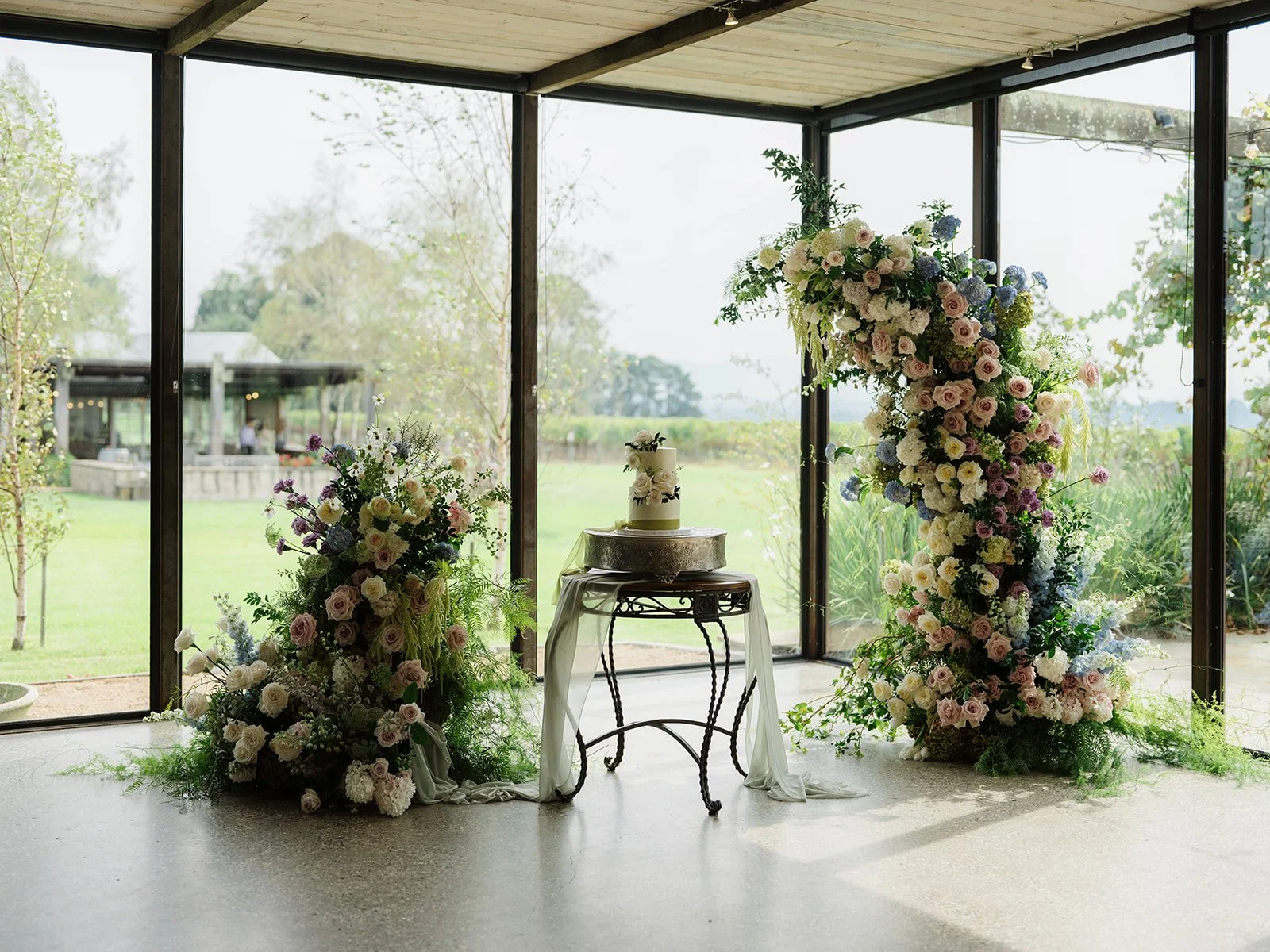 A wedding setup with floral arrangements, a cake on a stand, large windows with an outdoor view, and fairy lights.