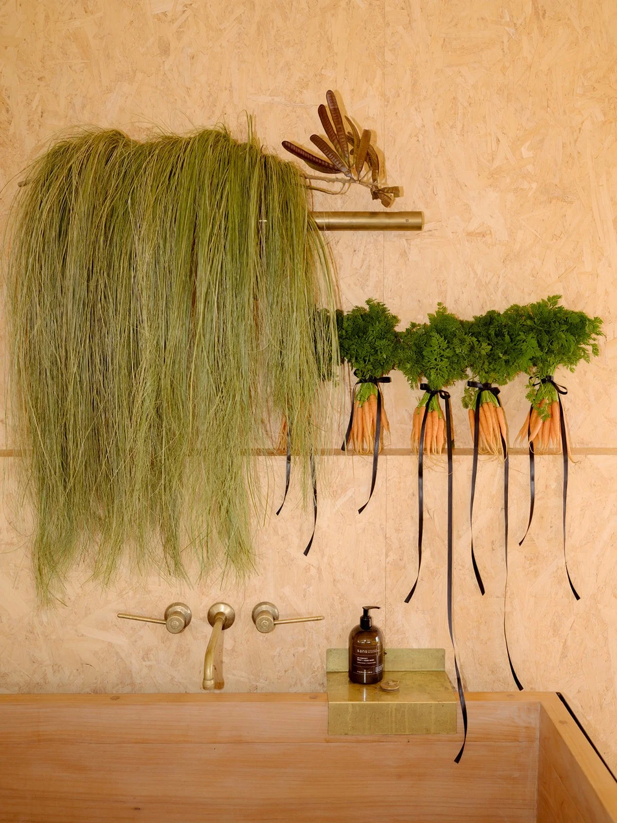 Fresh green herbs and vegetables hanging in a minimalist kitchen with wooden accents. A large bunch of green grass or herbal plant is hanging on a rod, and a row of carrot bundles tied with black ribbons is hanging below it. There is a soap dispenser