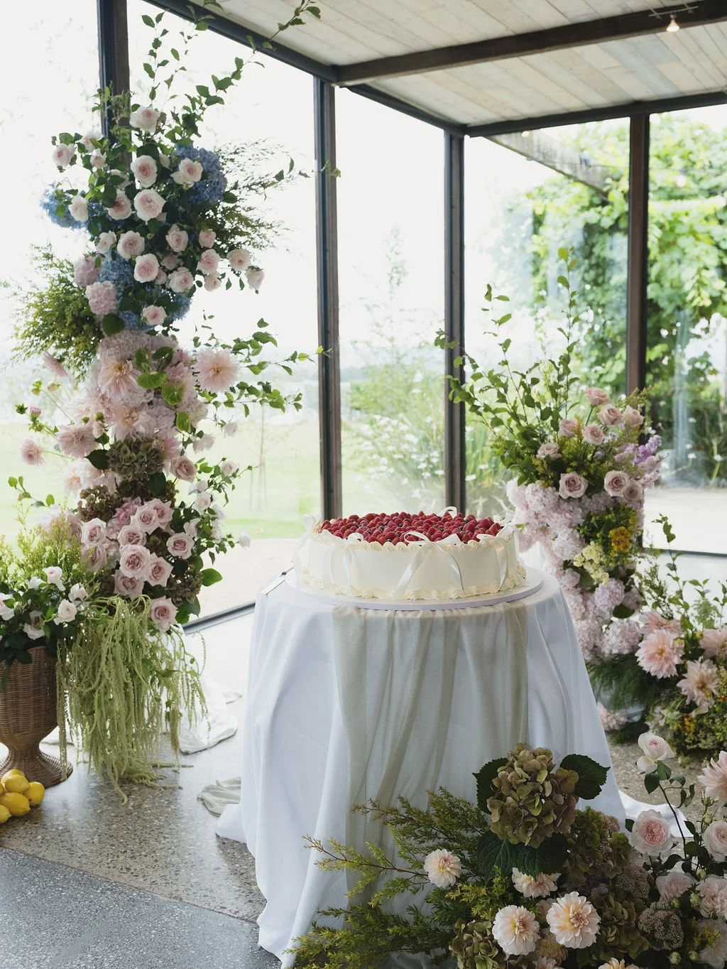 A white cake with strawberries on top on a table draped with white cloth, surrounded by pink and purple flowers in vases, inside a glass-enclosed space with greenery outside.