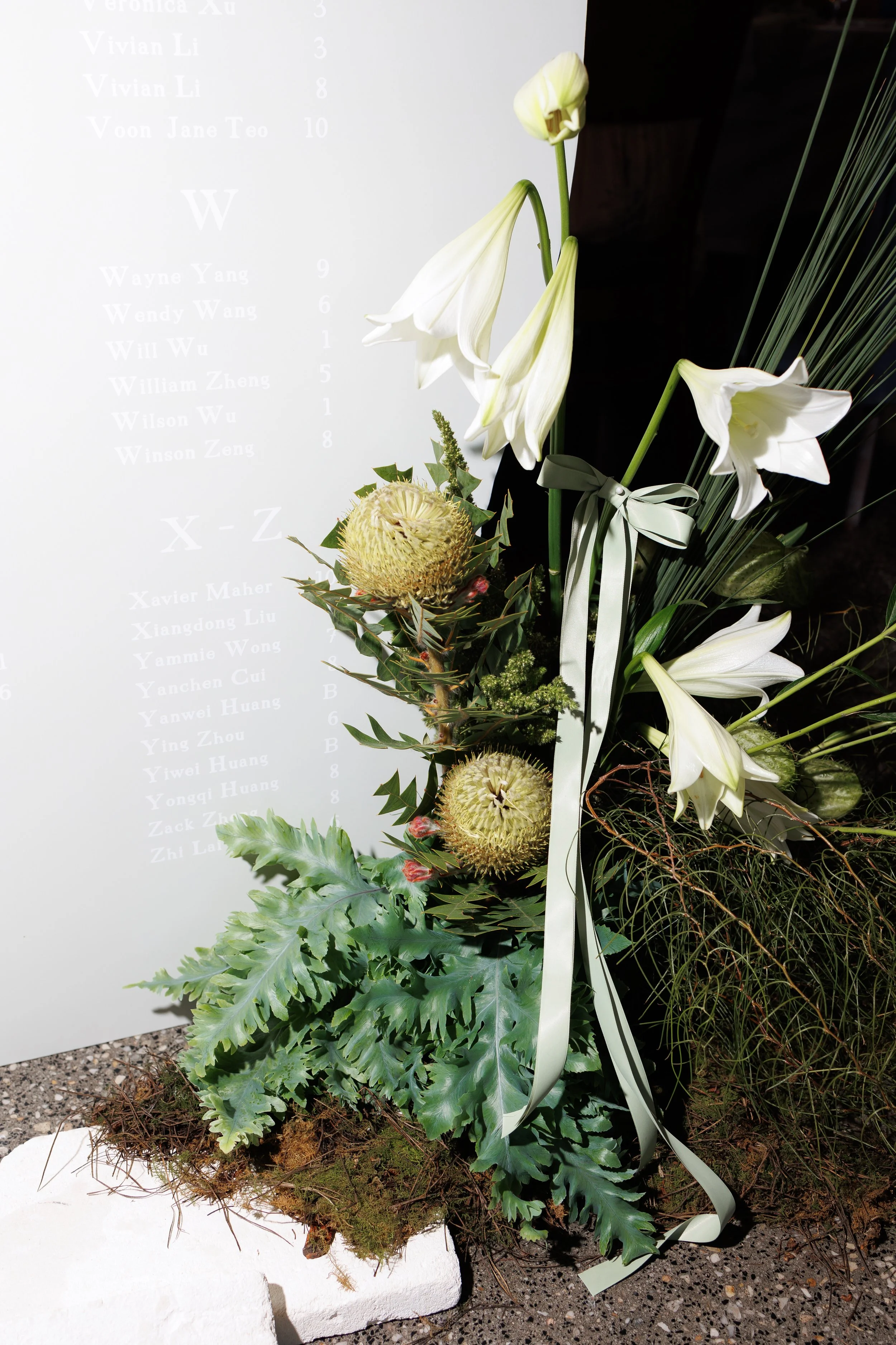A floral arrangement with white lilies, proteas, greenery, and a white ribbon, placed on a white platform in front of a board displaying names and numbers.