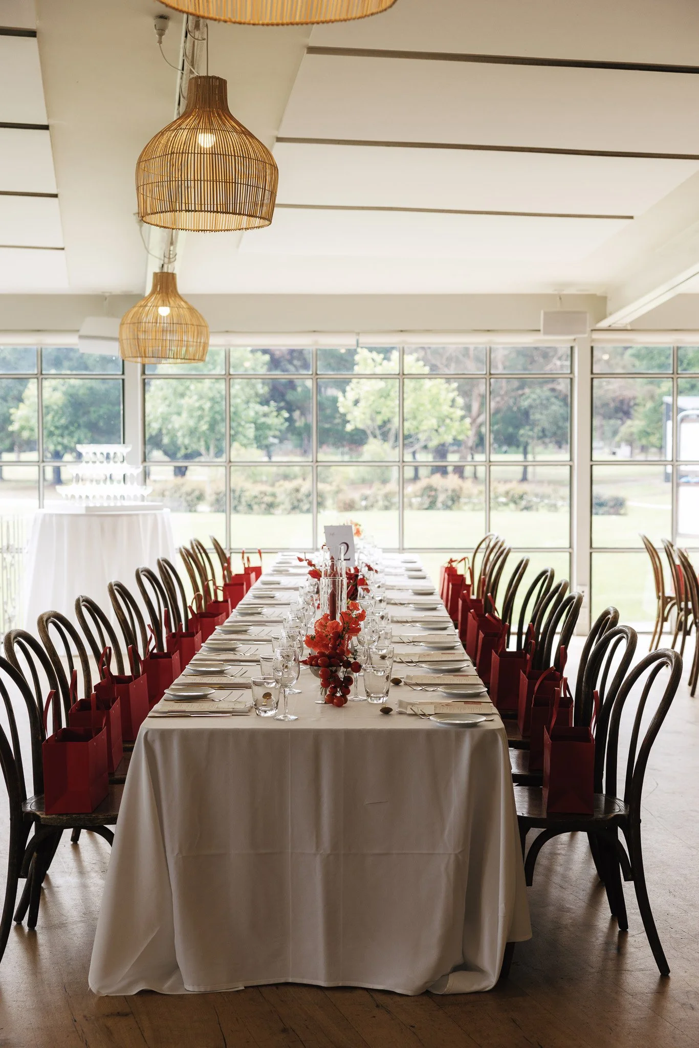 A long dining table set for a formal event with white tablecloth, glassware, and red gift bags on each seat, in a room with large windows and hanging wicker light fixtures.