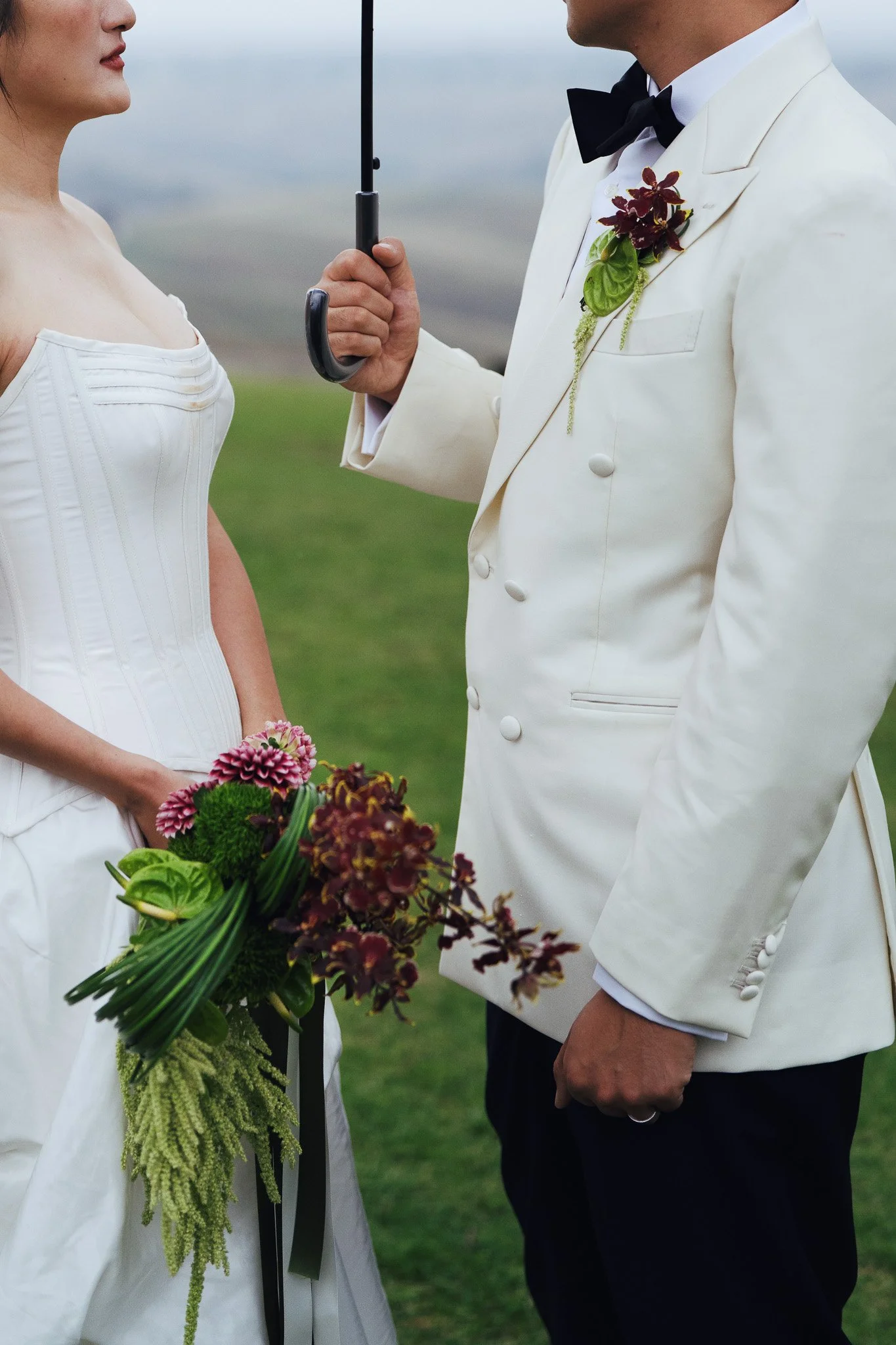 A bride and groom standing outdoors for their wedding ceremony, with the groom holding a black umbrella.