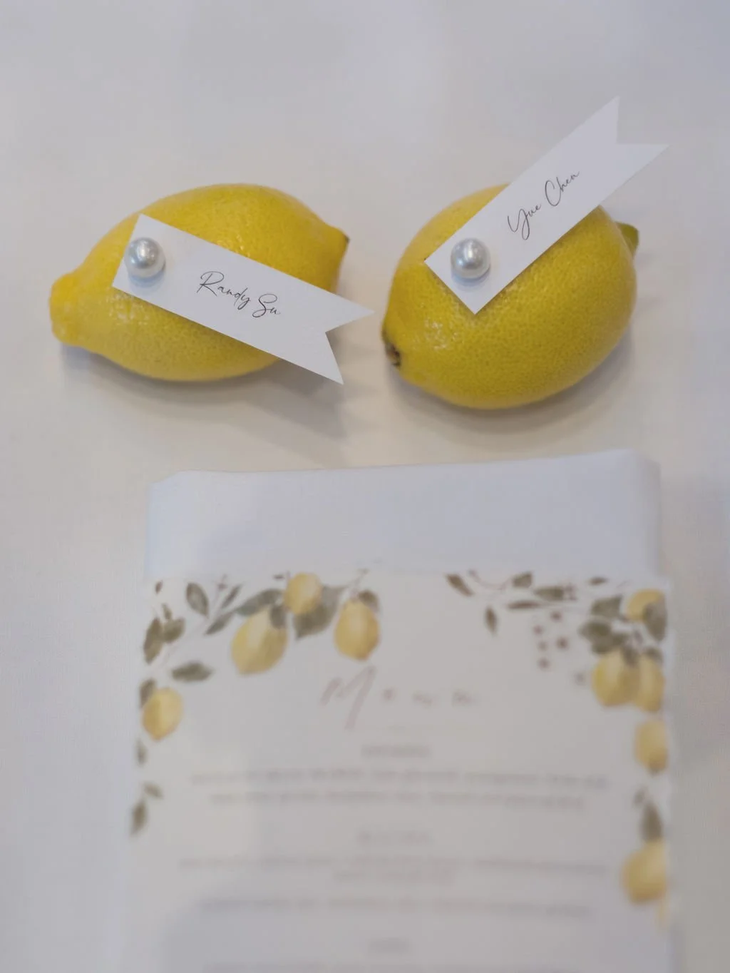 Two yellow lemons with white flags labeled 'Randy Sue' and 'You Charlotte', decorated with pearl pins, on a white surface in front of a blurred box featuring lemon illustrations.