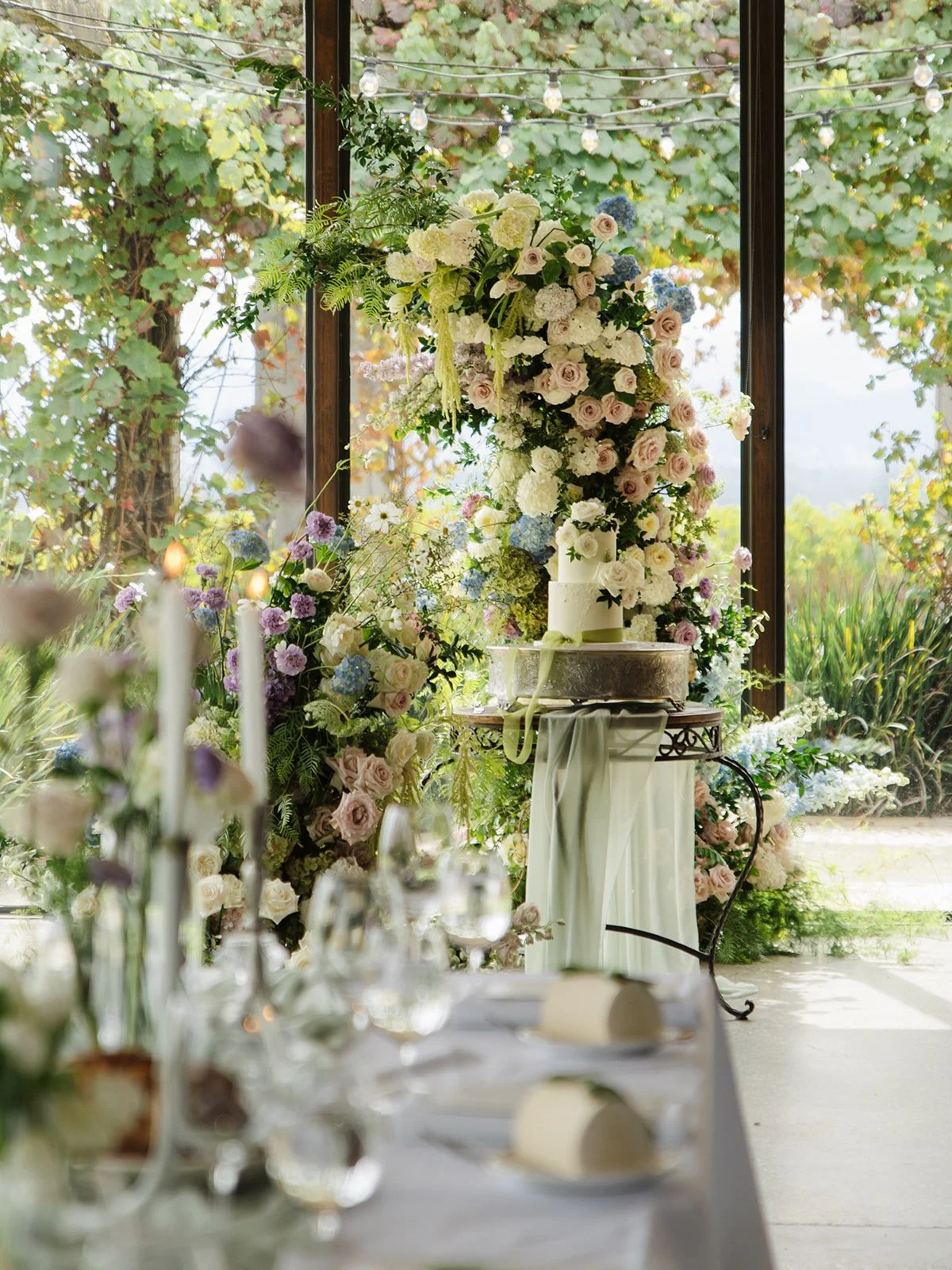Elegant wedding cake with white flowers at front, decorated with a ribbon, on a table surrounded by floral arrangements, with a window and outdoor greenery in the background.