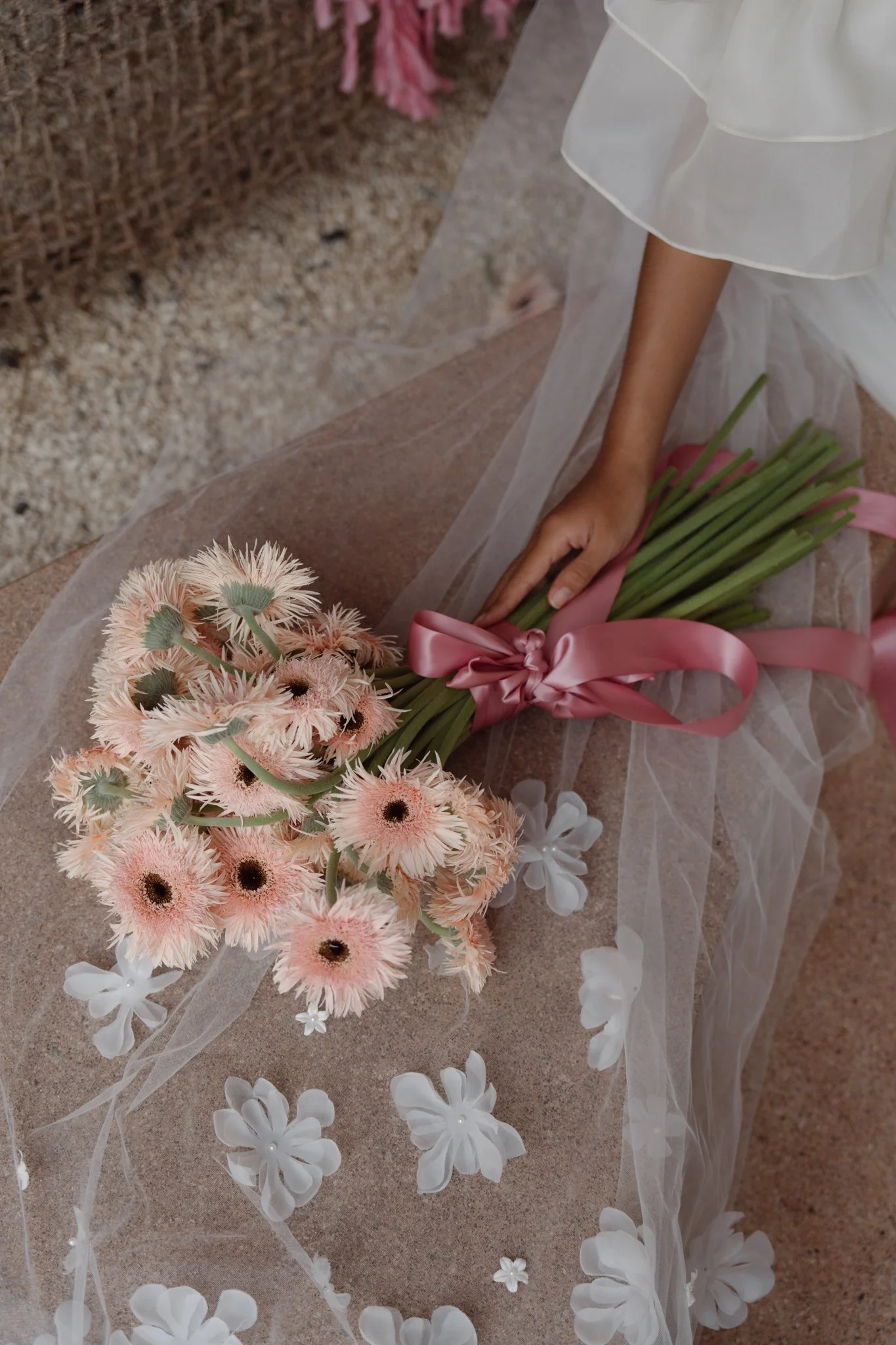 A person wearing a white dress is setting a bouquet of pink flowers on a surface covered with white floral and sheer fabric decoration. The bouquet is tied with a pink ribbon.