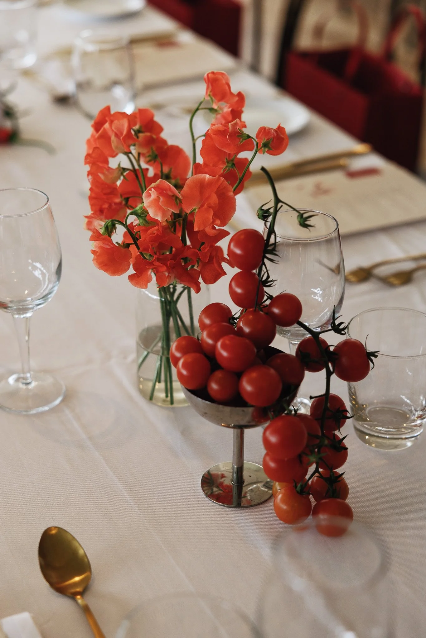 A table decorated with a centerpiece of orange flowers and a string of cherry tomatoes, surrounded by wine glasses and gold utensils in a formal dining setting.